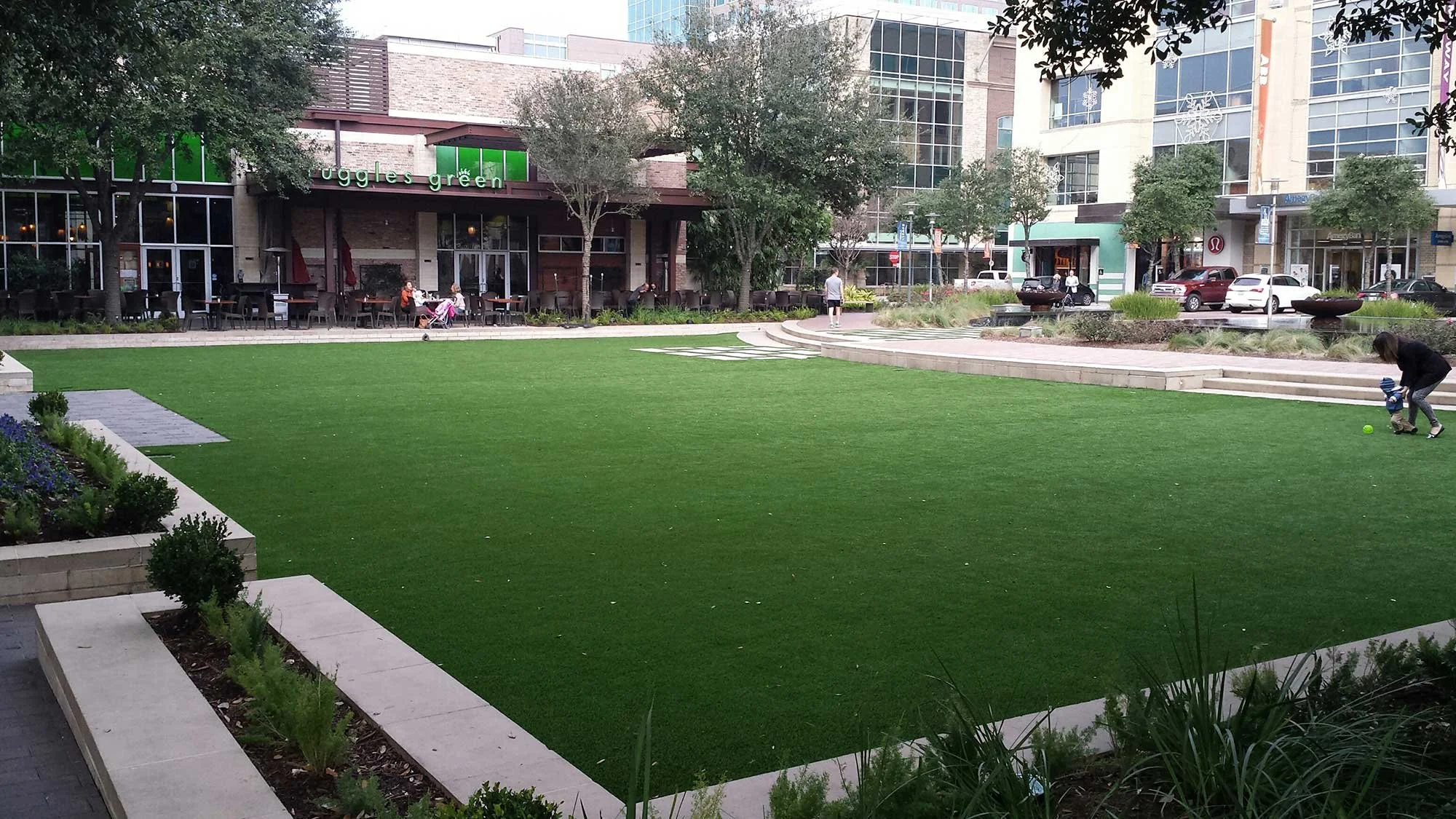 Urban park with a vibrant green artificial grass area, surrounded by steps and garden beds. People are seen sitting at a restaurant and walking, with a child playing with a ball in the foreground. Modern buildings with glass windows and a store with a green sign are in the background.