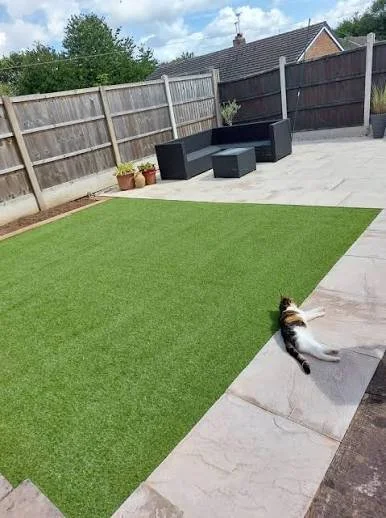 Backyard with artificial grass area, potted plants, outdoor black furniture, and a cat lying on the stone patio.
