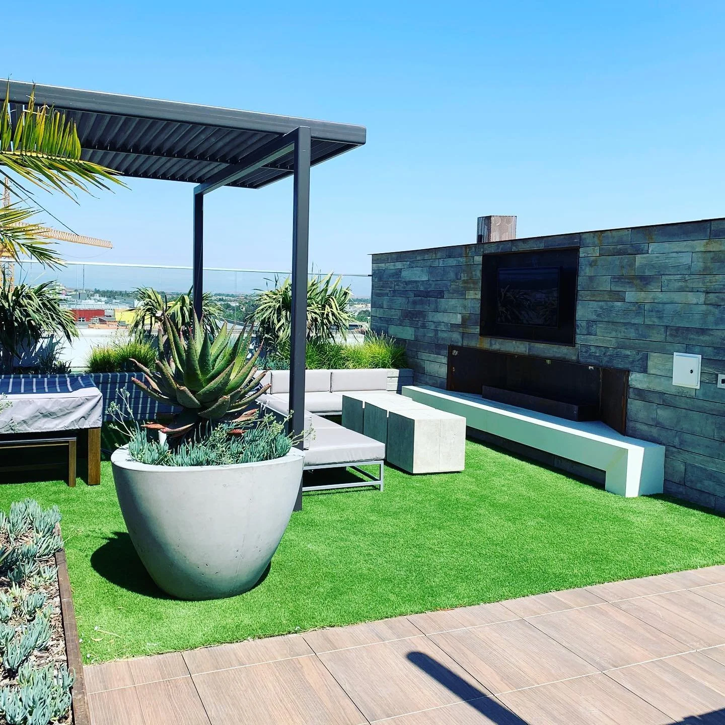 Rooftop terrace with green artificial grass, large potted succulents, modern seating, and a wall-mounted television under a canopy, city skyline in the background