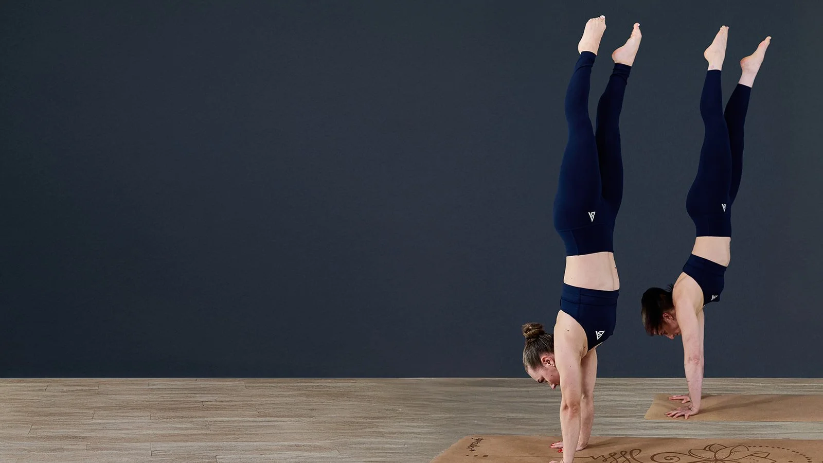 Zwei Frauen machen Yoga im Handstand in einem Raum mit dunklem Hintergrund und Holzboden.