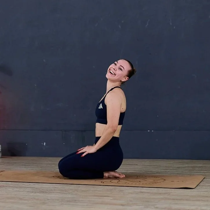 Frau in Sportbekleidung, die auf einem Yogamatte in einer Turnhalle sitzt, lächelnd.