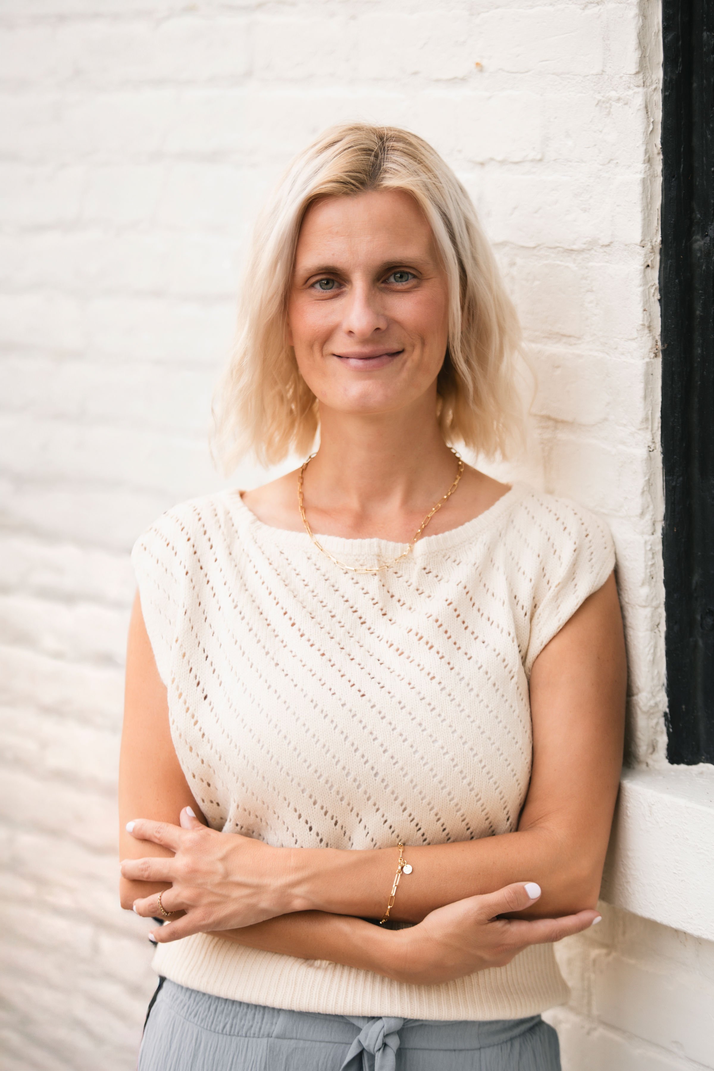 A woman with blonde hair standing against a white brick wall, wearing a cream-colored knit top and gray drawstring pants, smiling with folded arms.