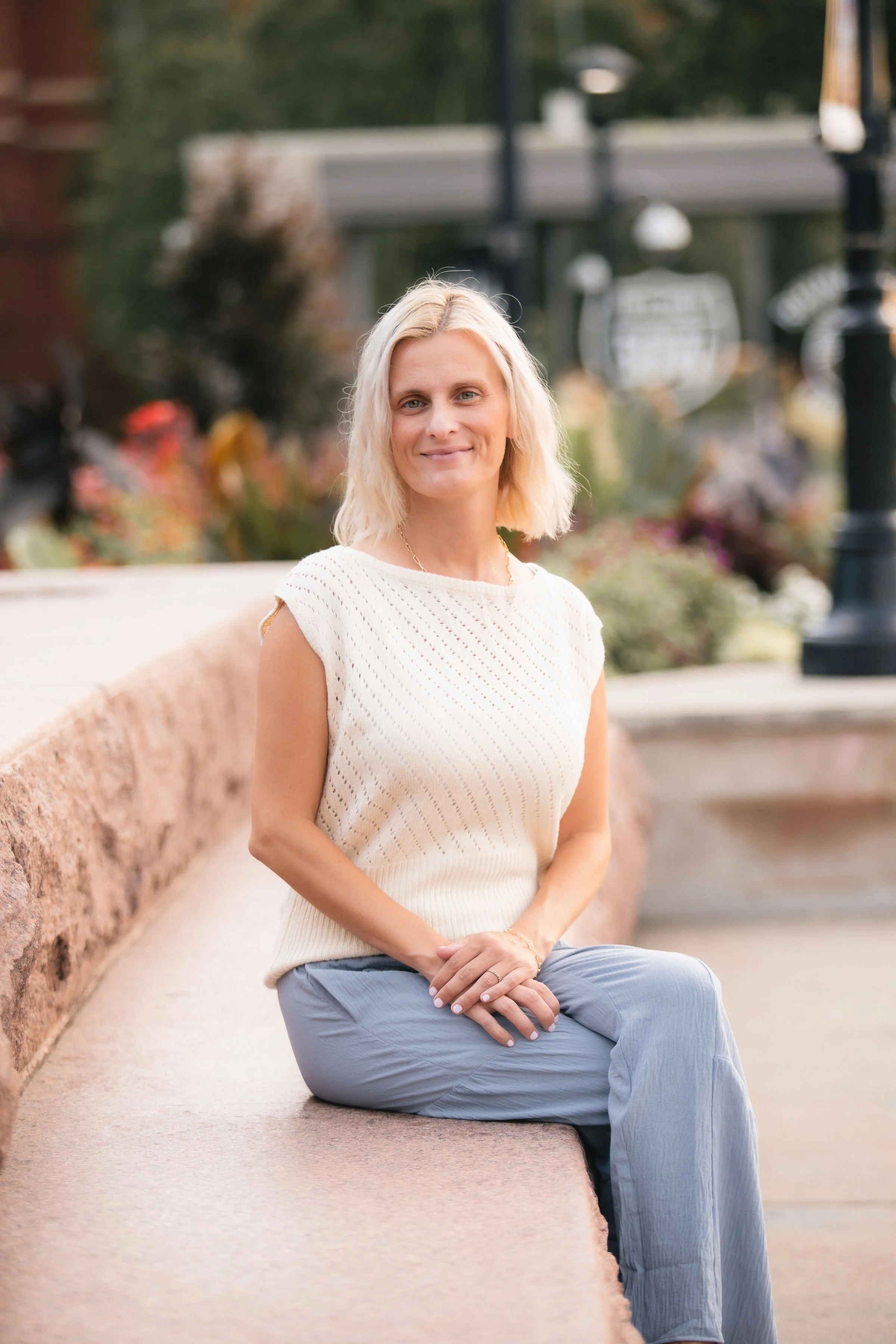 A woman with blonde hair, wearing a white sleeveless top and blue pants, sitting on a pinkish stone bench outdoors with a background of greenery and black lamp posts.