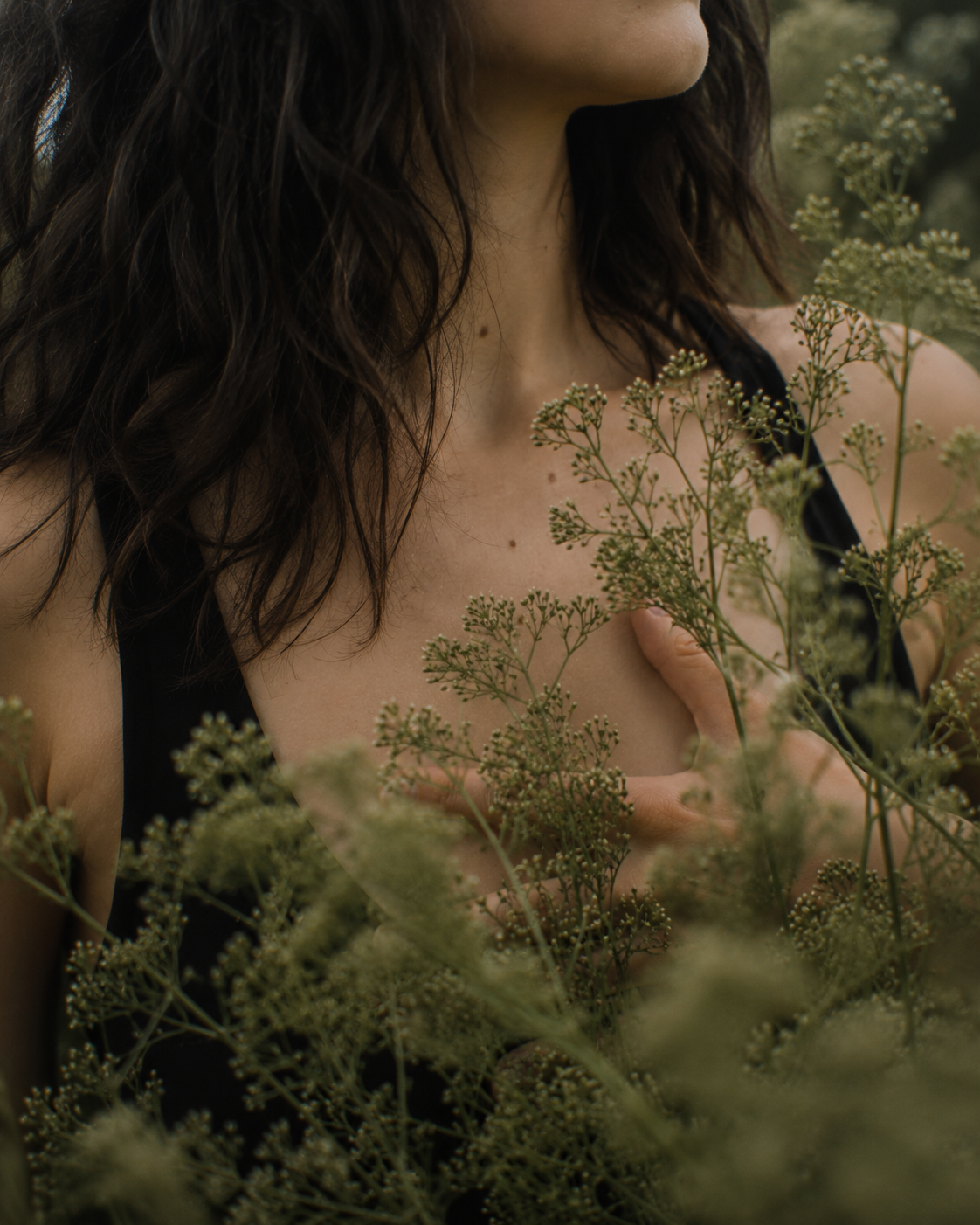A woman with dark, curly hair wearing a black tank top, standing amidst green plants, covering her chest with her hand.