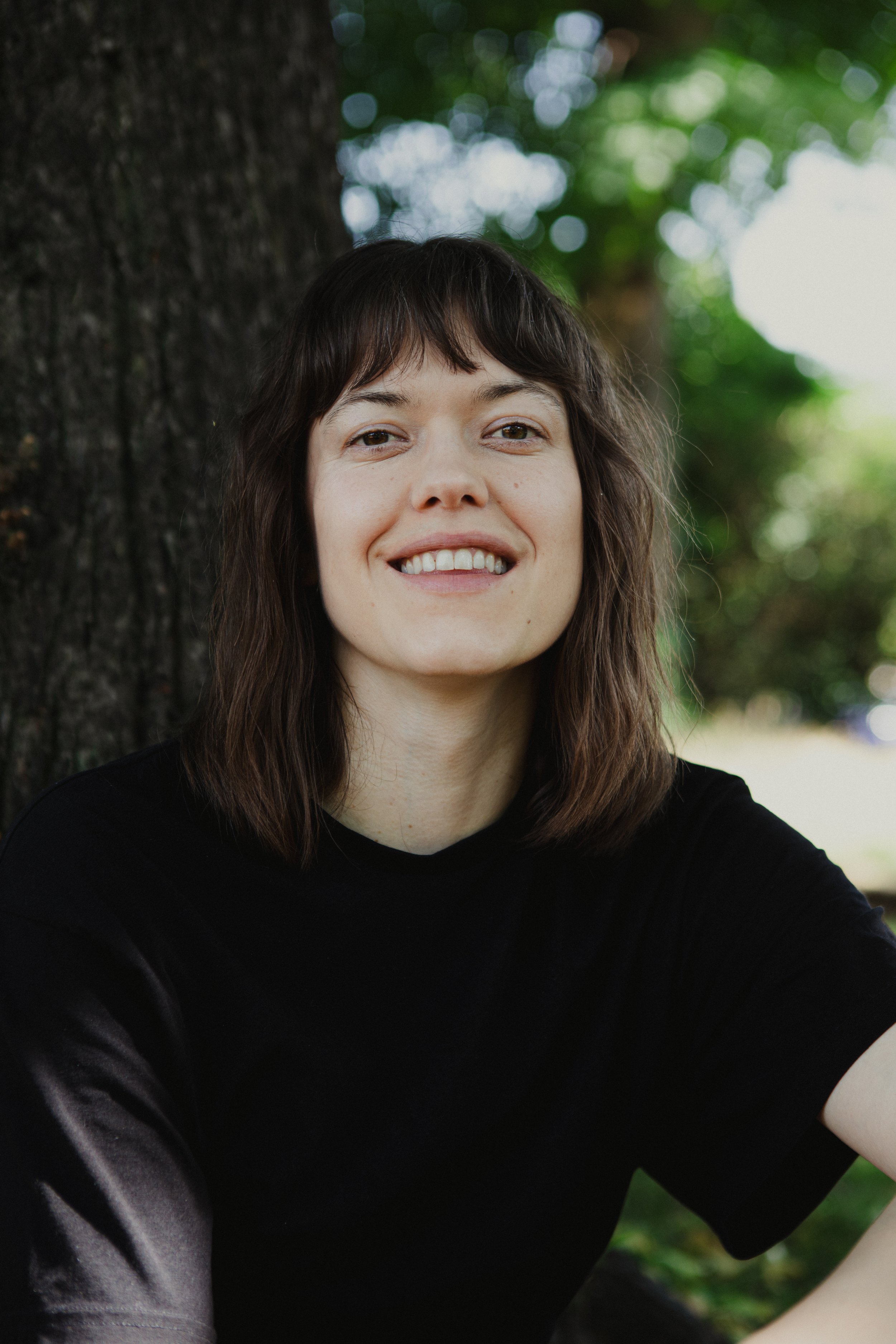 A young woman with shoulder-length brown hair smiling outdoors with a tree trunk and green foliage in the background.