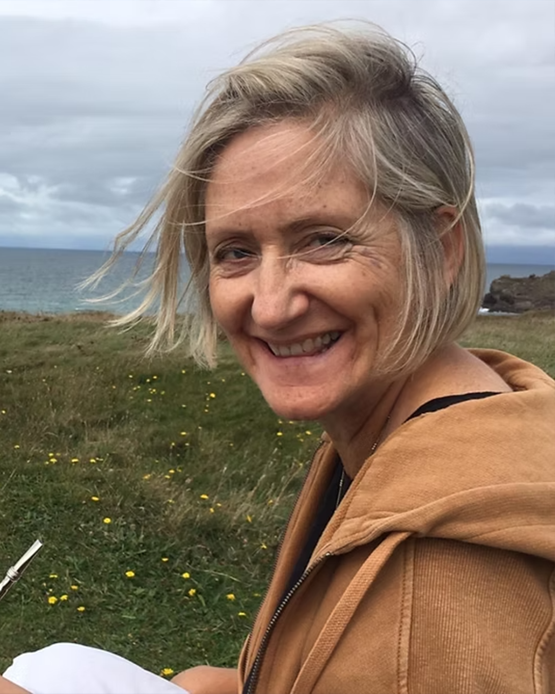 A woman smiling outdoors near the beach during cloudy weather, with wind-blown short blonde hair and a light brown jacket.