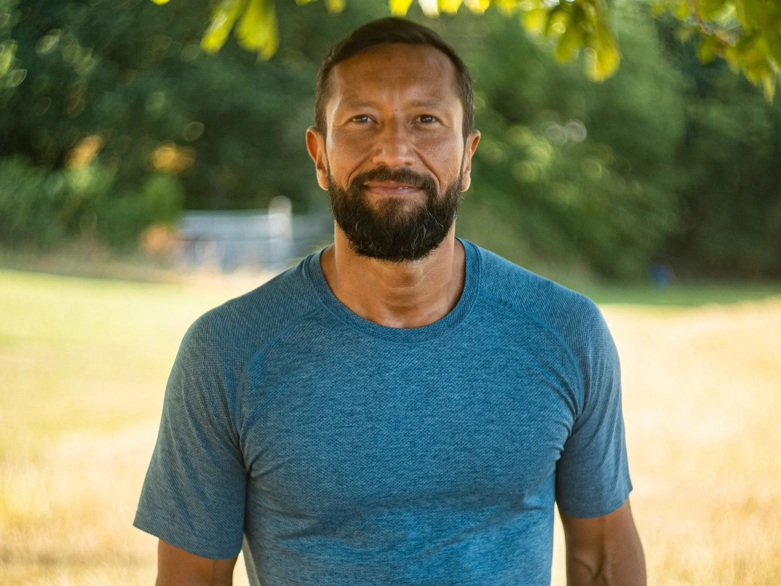 A man with a beard and short hair smiling outdoors in a green park, wearing a blue athletic shirt.
