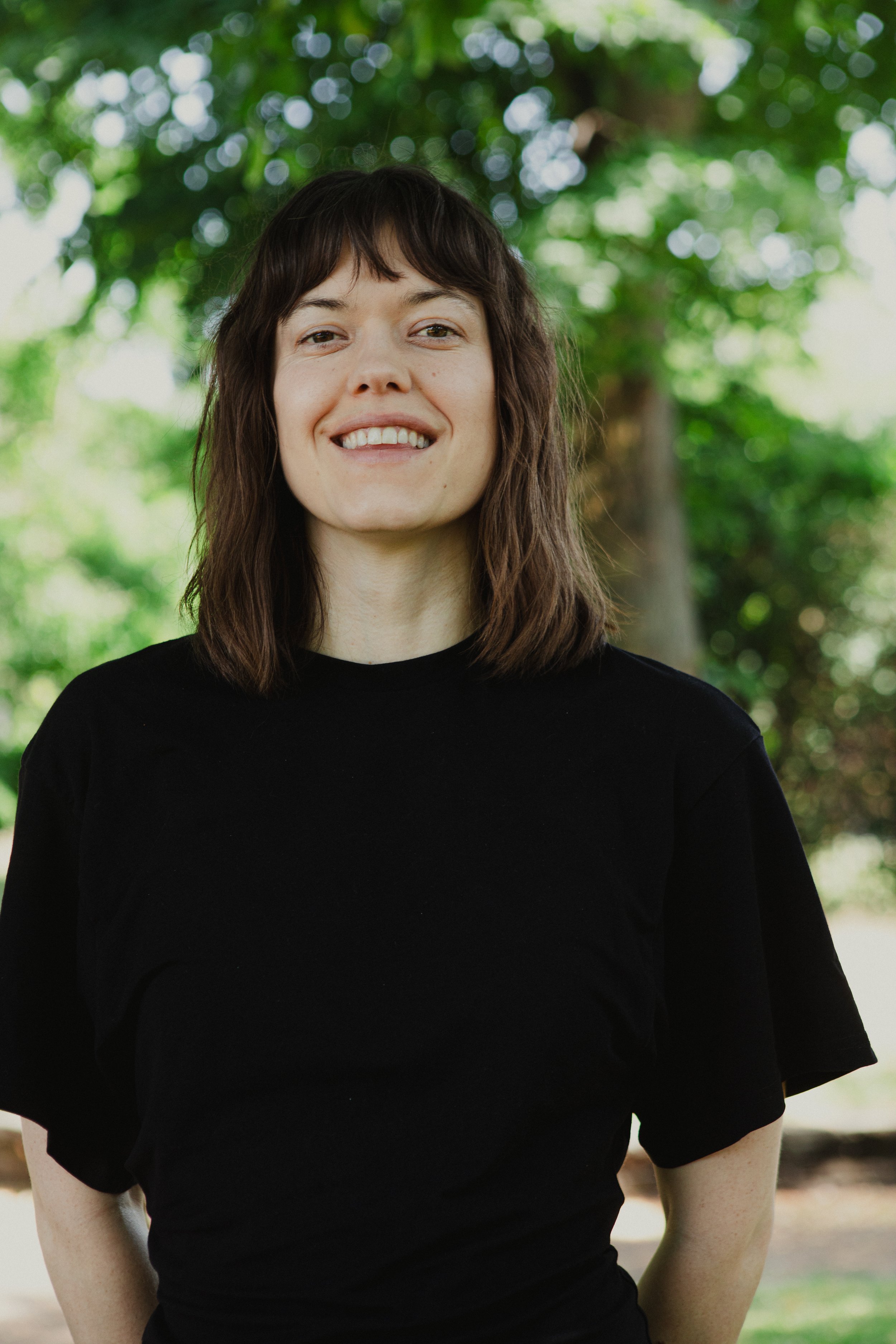A woman with shoulder-length brown hair, wearing a black shirt, smiling outdoors with greenery and blurred trees in the background.