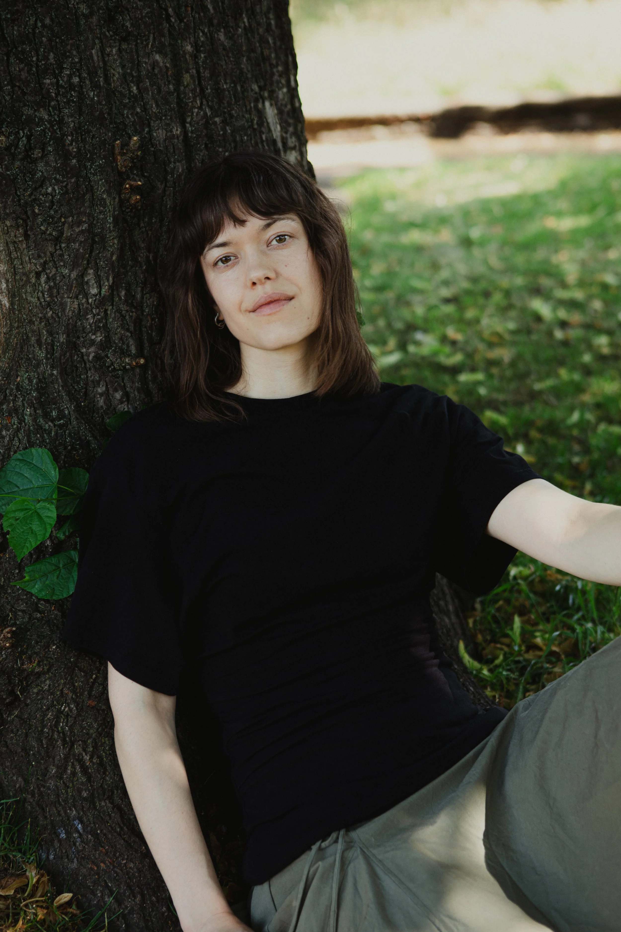 A young woman with shoulder-length brown hair and fair skin sits leaning against a tree trunk outdoors. She wears a black T-shirt and gray pants, and looks at the camera with a calm expression. The background shows green grass and blurred foliage.