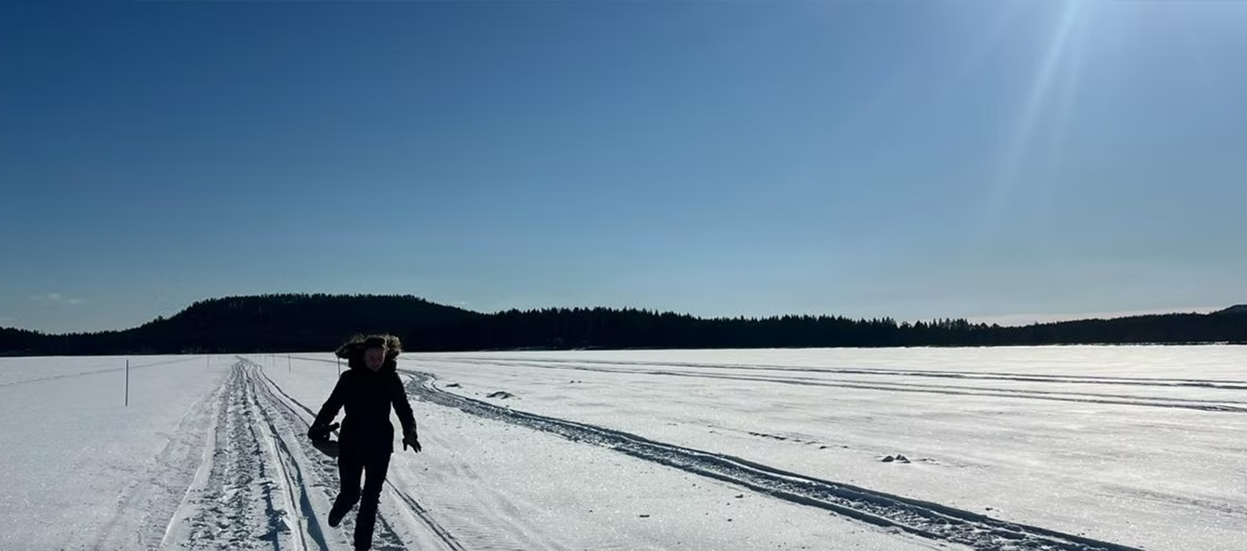 A person walking on snowy tracks in a vast, snow-covered landscape with a forested hill in the background under a clear blue sky.