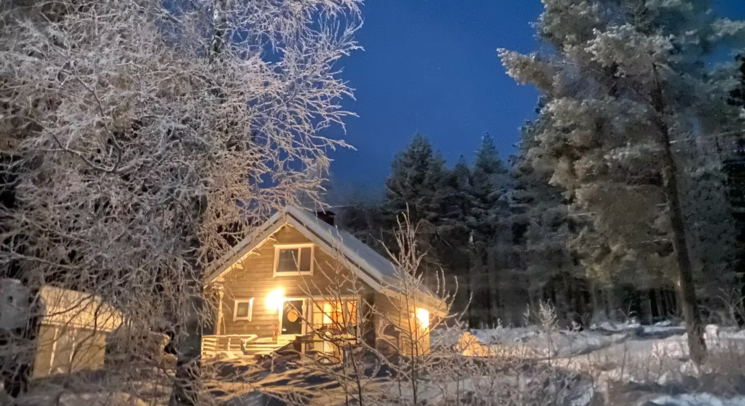 A snow-covered house with a lit porch light, surrounded by snow-covered trees under a dark blue evening sky.