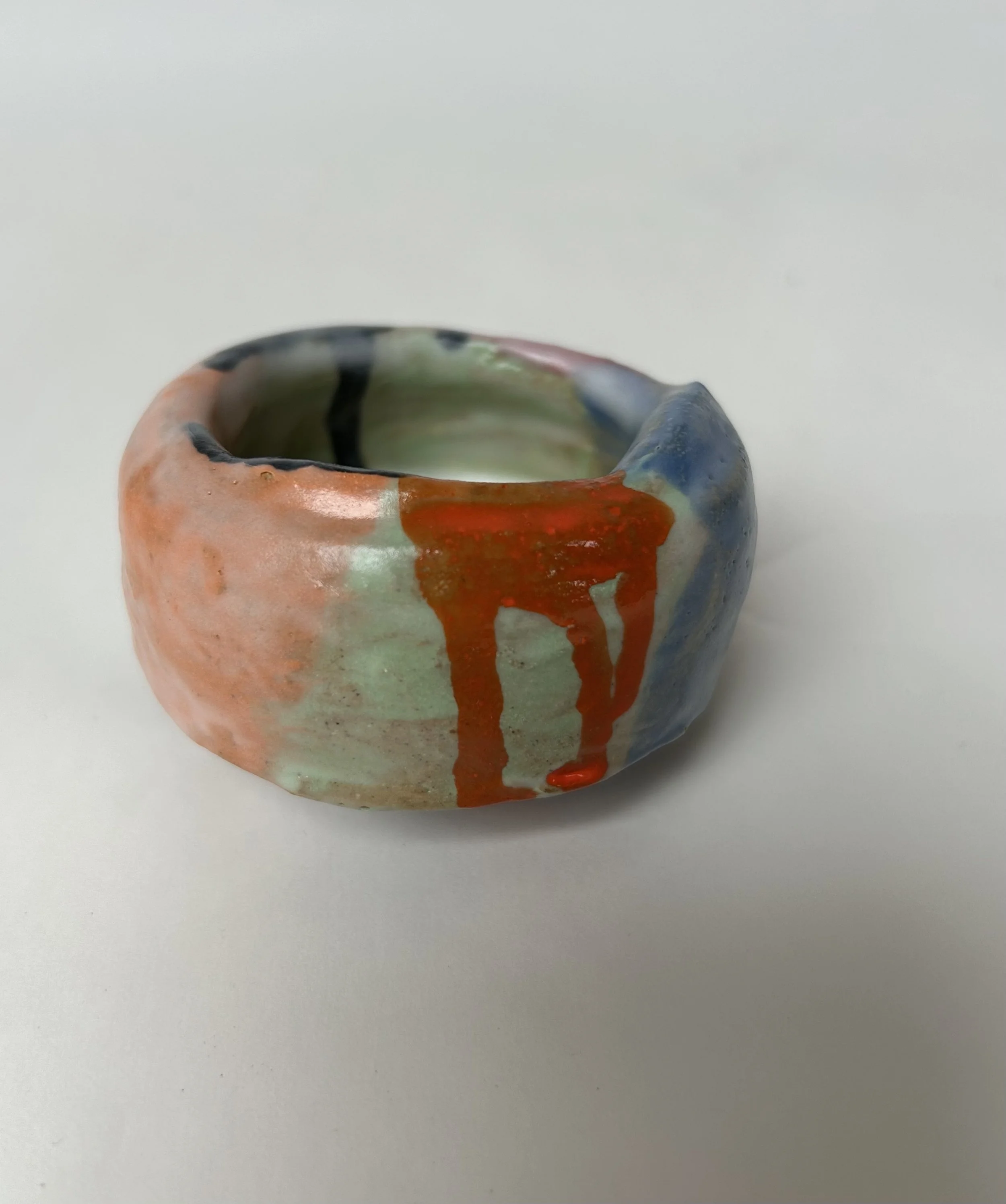 A small, handmade ceramic cup with multicolored glaze, featuring orange, blue, green, and black accents, placed on a plain white surface.