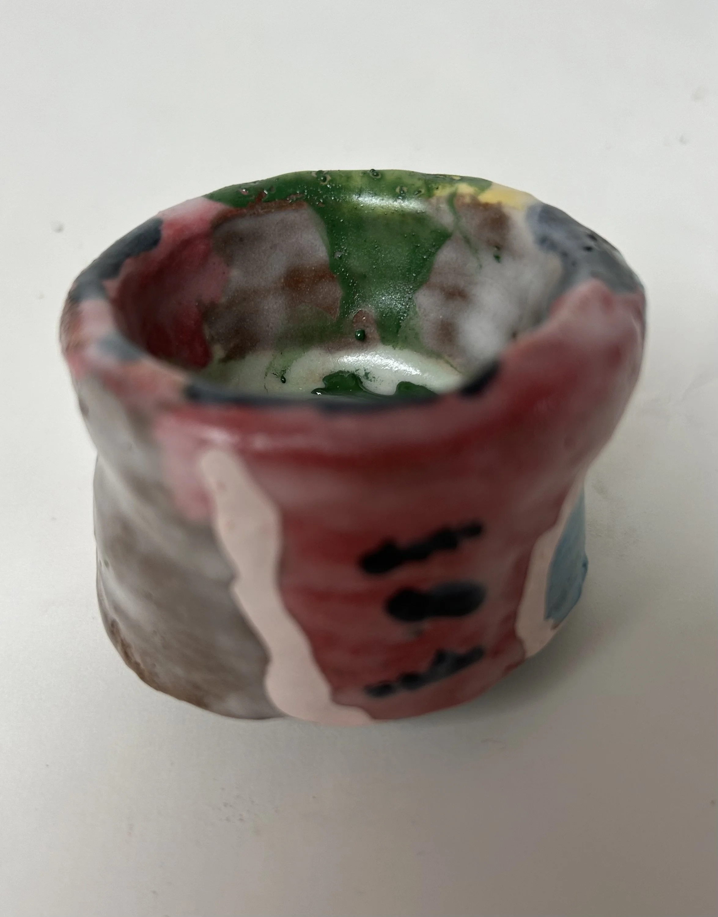 Close-up of a small, colorful ceramic or paper mache bowl with a cloudy, marbled pattern of pink, gray, green, and black, placed on a plain white surface.