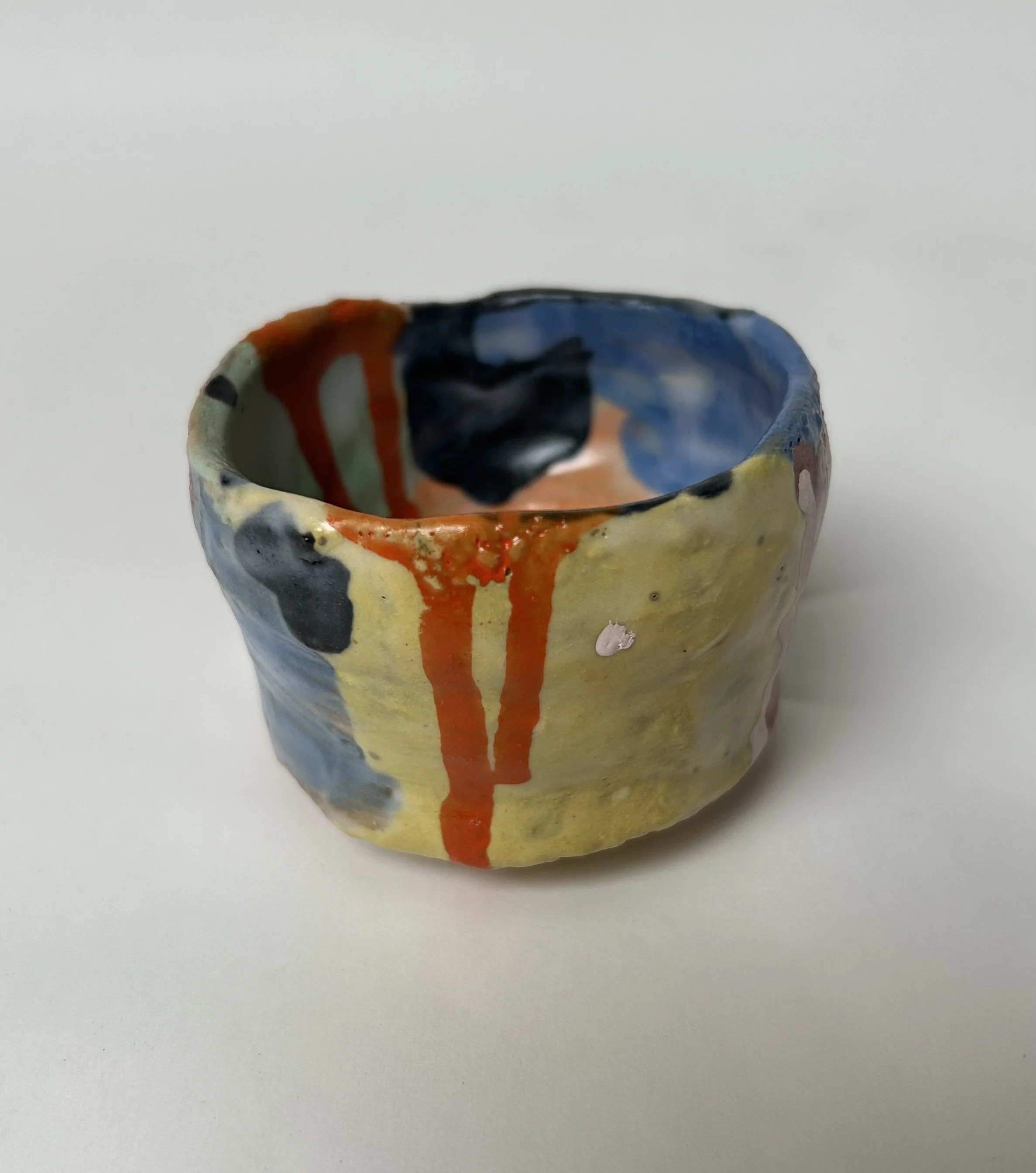 A small, colorful, ceramic cup with a rounded shape, painted with abstract patterns in shades of blue, orange, black, and white, placed on a plain white surface.