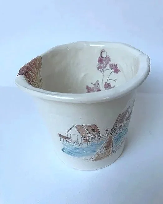 A decorative ceramic cup with a hand-painted scene of a house by water, trees, and a small boat.