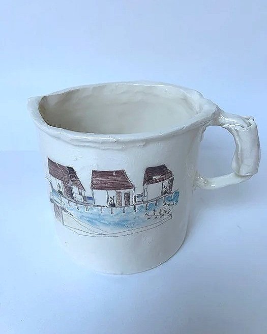 A white ceramic mug with a beach hut illustration and a blue water design, featuring a handle on the right side, on a light background.