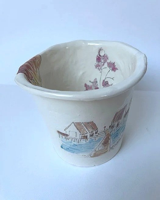 Empty ceramic cup with a painted rural scene, including houses, a dock, and trees.