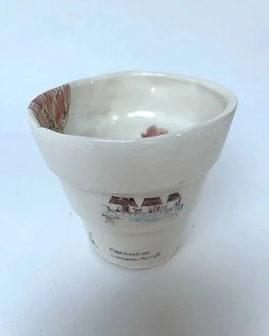 A white ceramic cup with a curved shape, decorated with an image of a beach scene with huts and palm trees, placed on a white surface.