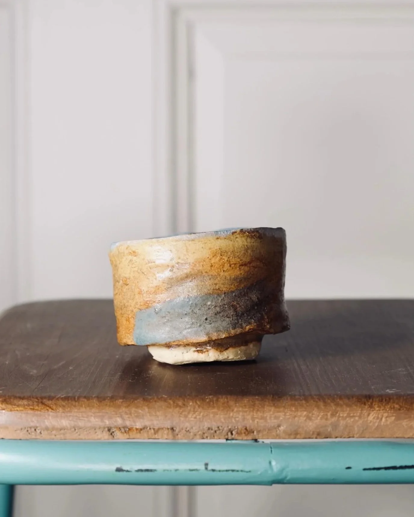A ceramic cup with a rustic, glazed exterior, placed on a wooden surface. The background includes a light-colored wall with a door.