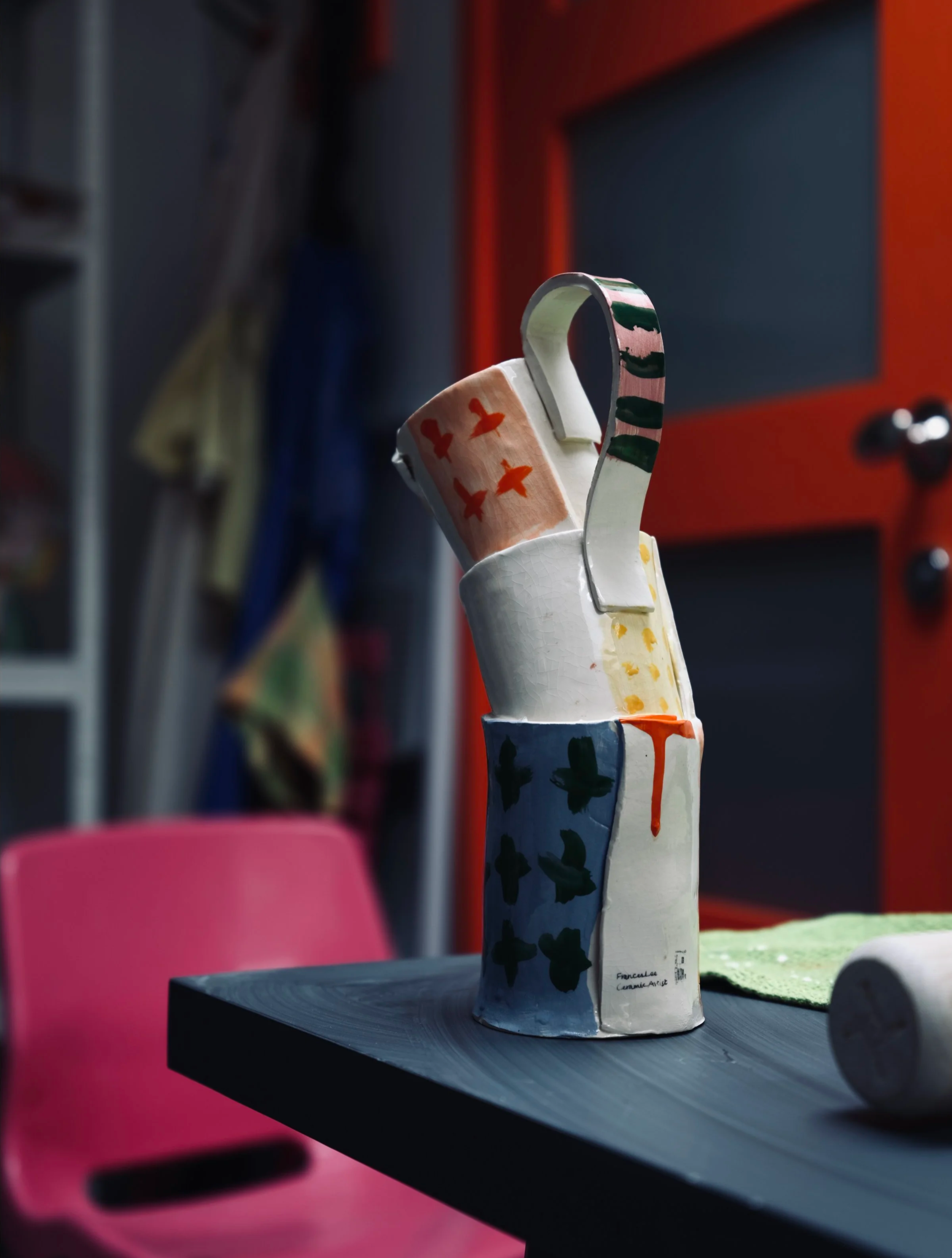 A ceramic sculpture of a hand with painted designs, placed on a black paper record on a dark table. The background features a blurred red and gray drawer and a pink chair.