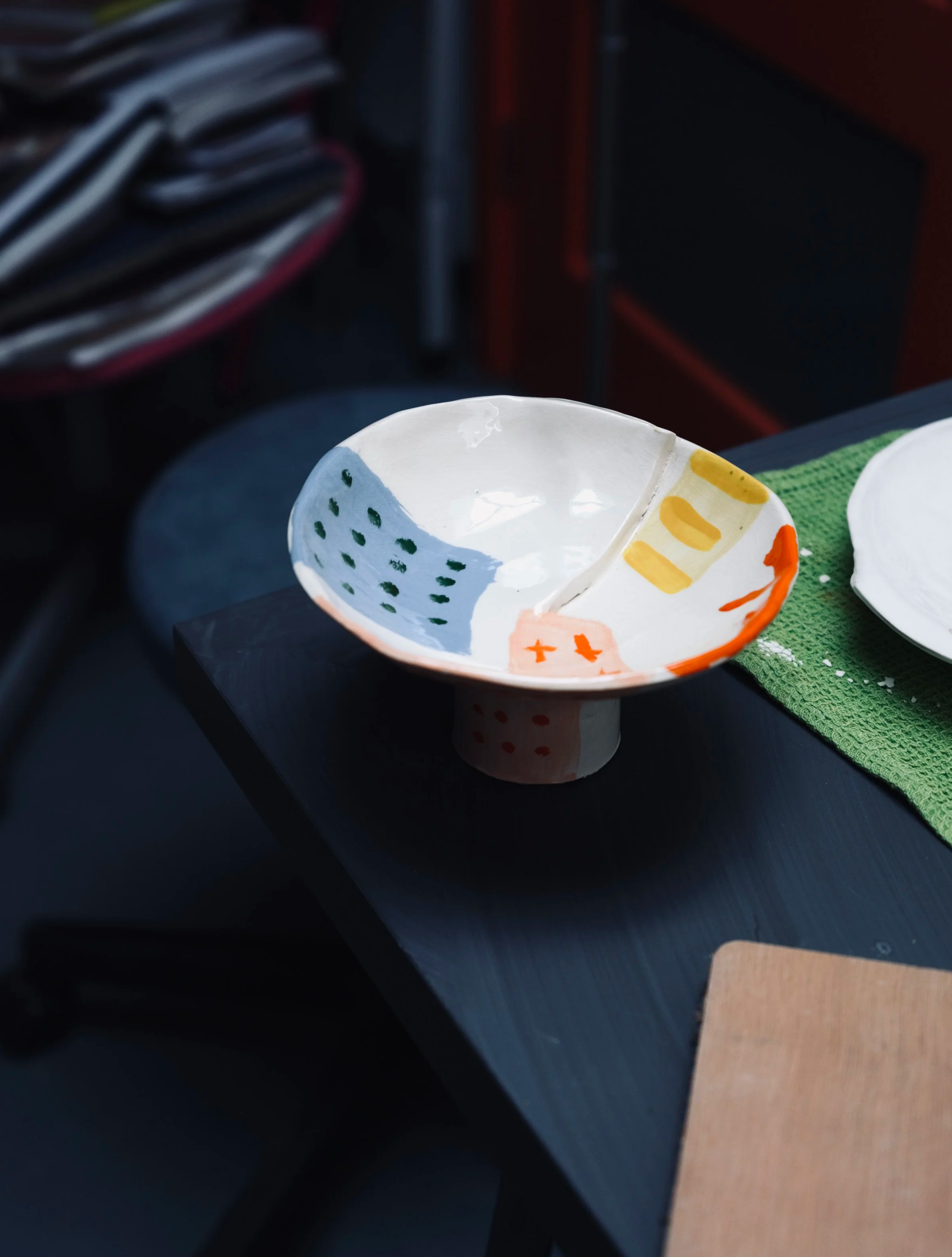 A ceramic bowl with colorful hand-painted patterns, placed on a dark surface next to a green textured cloth and a cardboard piece, with blurred background objects.
