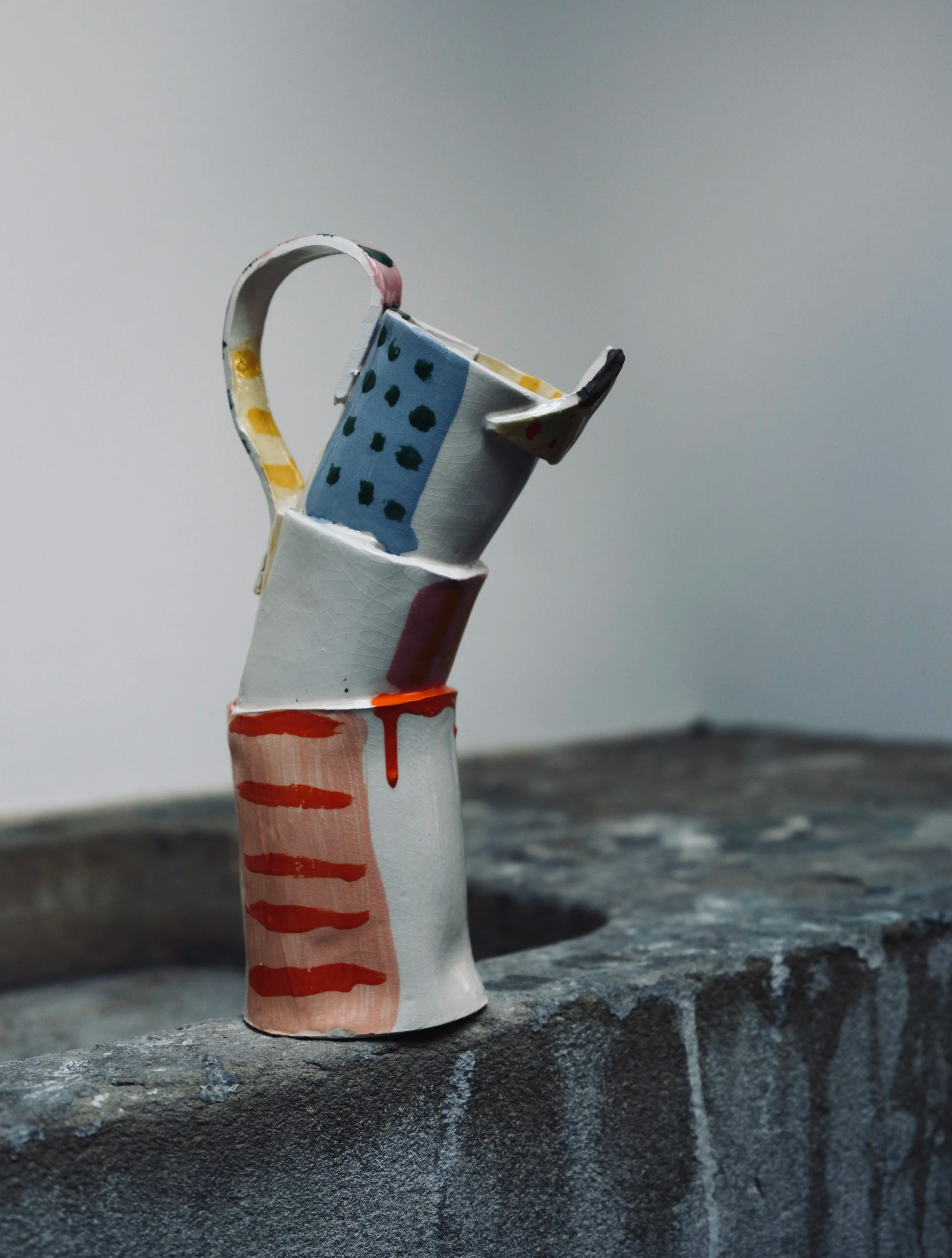 Close-up of a painted paper pitcher with a handle, decorated with polka dots and stripes, placed on a textured surface against a plain background.