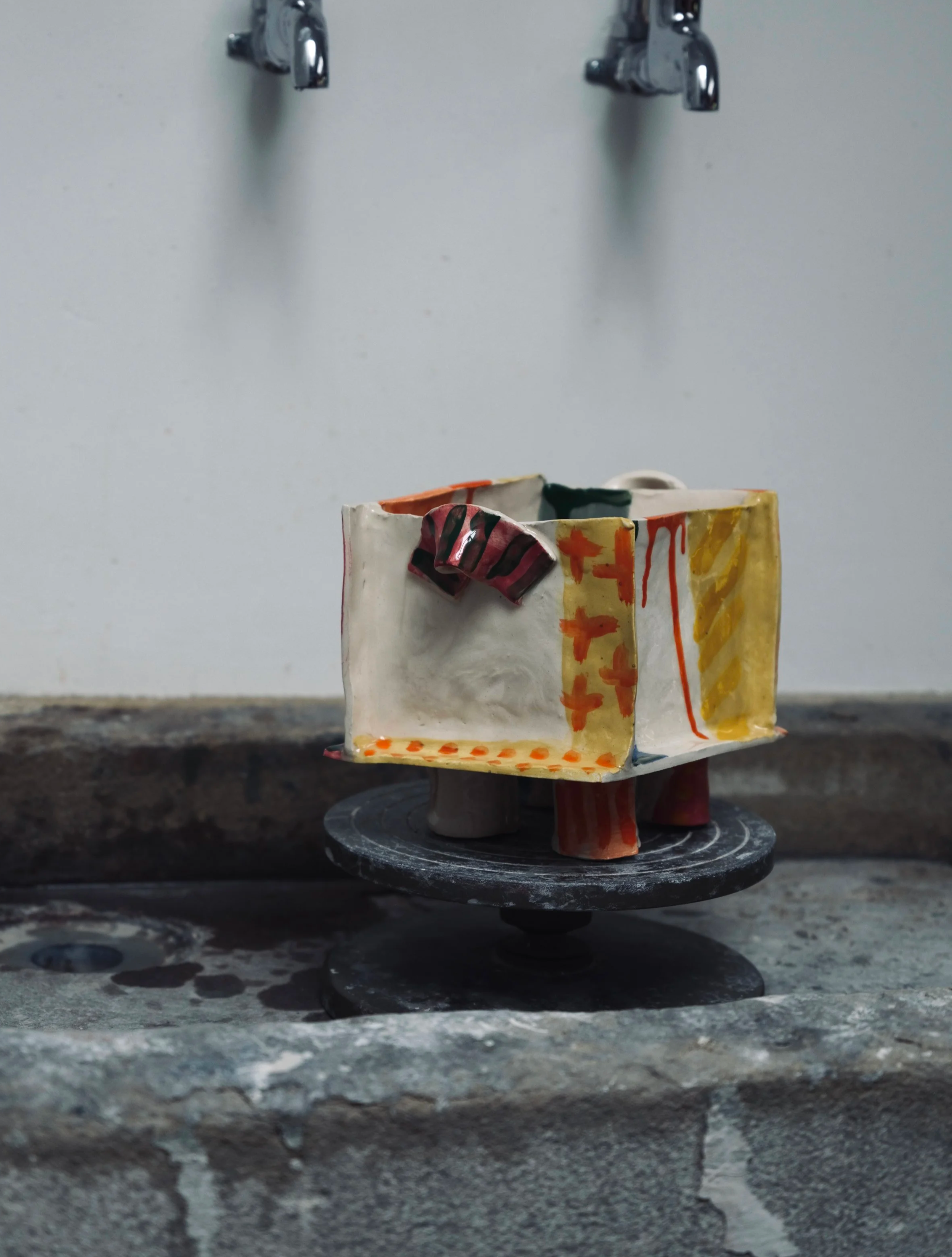 A small ceramic sculpture of a house with colorful abstract patterns resting on a black circular base, with the background featuring a plain white wall and two wall-mounted faucet handles.