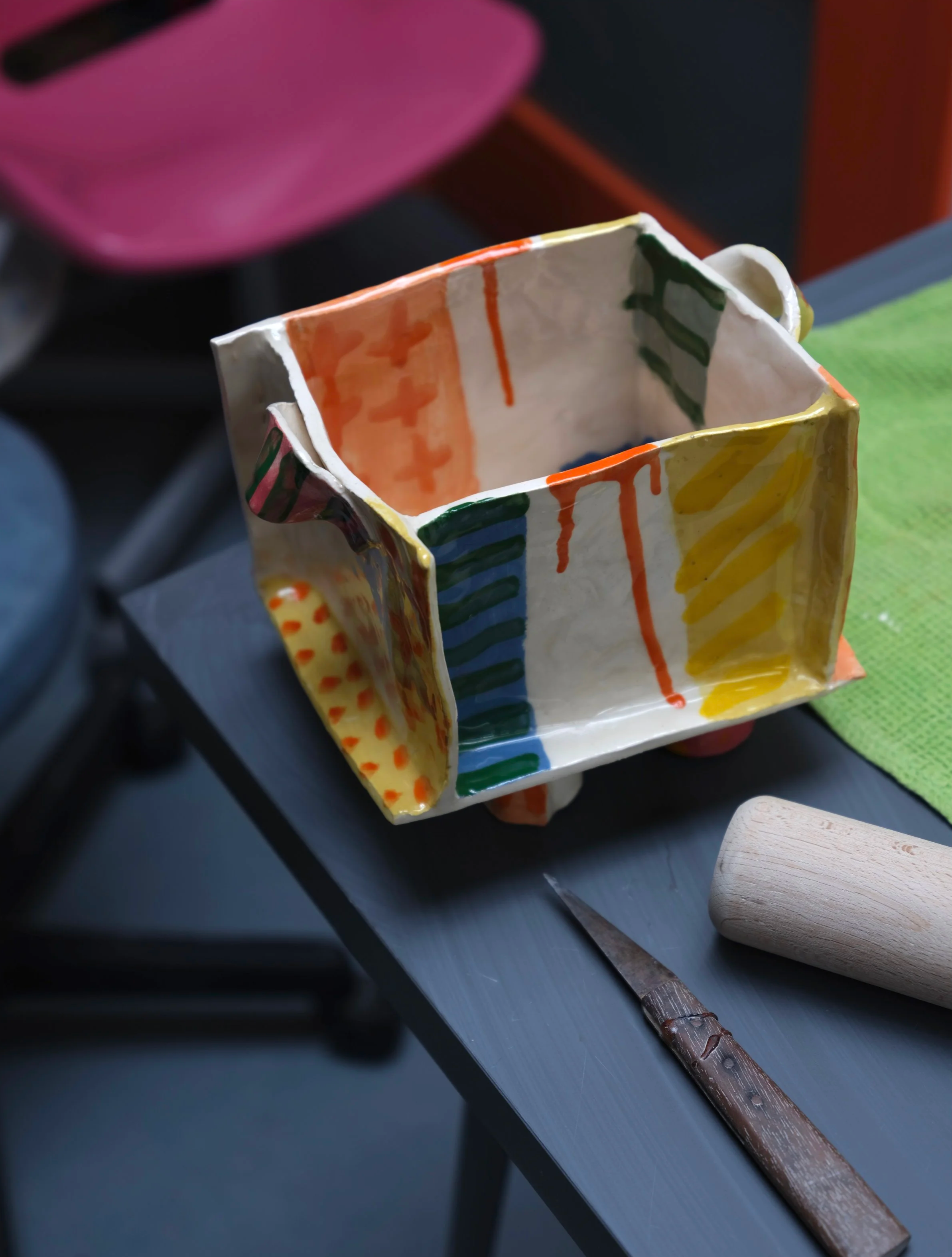 A colorful, handmade ceramic mug with painted stripes in orange, green, and yellow, sitting on a table near a green cloth and a chisel.