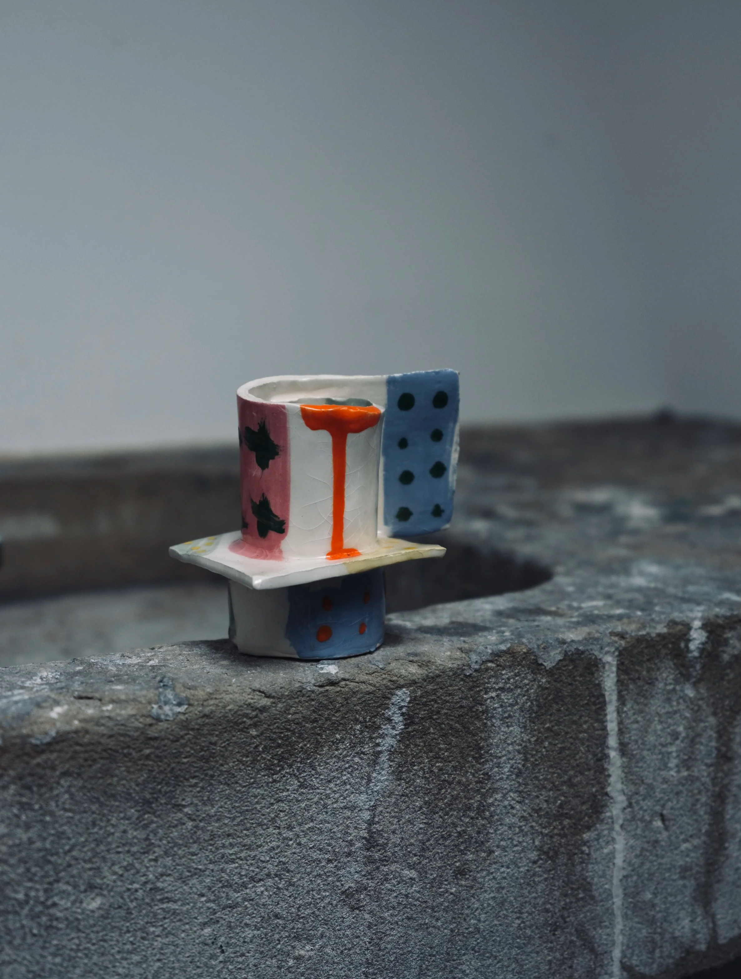 A small, hand-painted paper or clay sculpture resembling a person, standing on a rough stone surface, with a plain gray background.