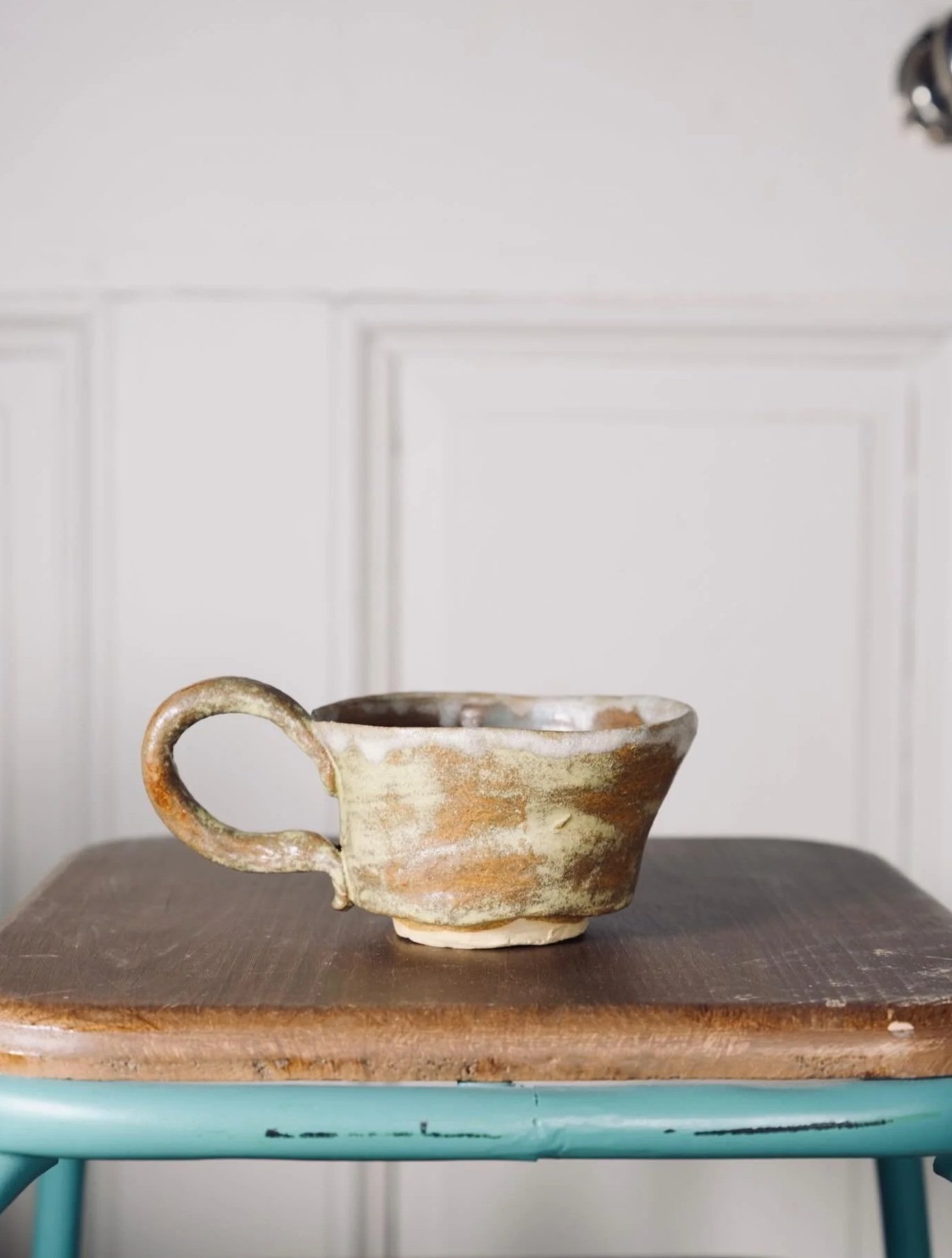 A ceramic cup with a handle, glazed in earthy tones, sitting on a wooden surface in front of a white paneled wall.