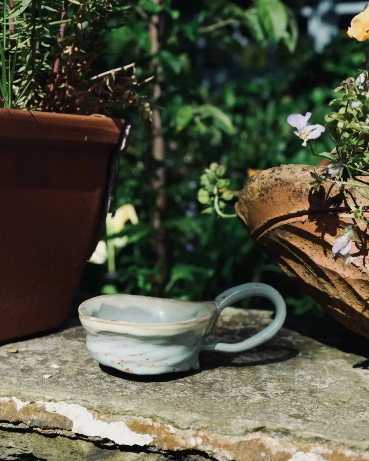 A small white ceramic cup with a handle and warm glaze, placed on a rough stone surface outdoors. Surrounding elements include a terra cotta flowerpot, green foliage, and white flowers on the right.