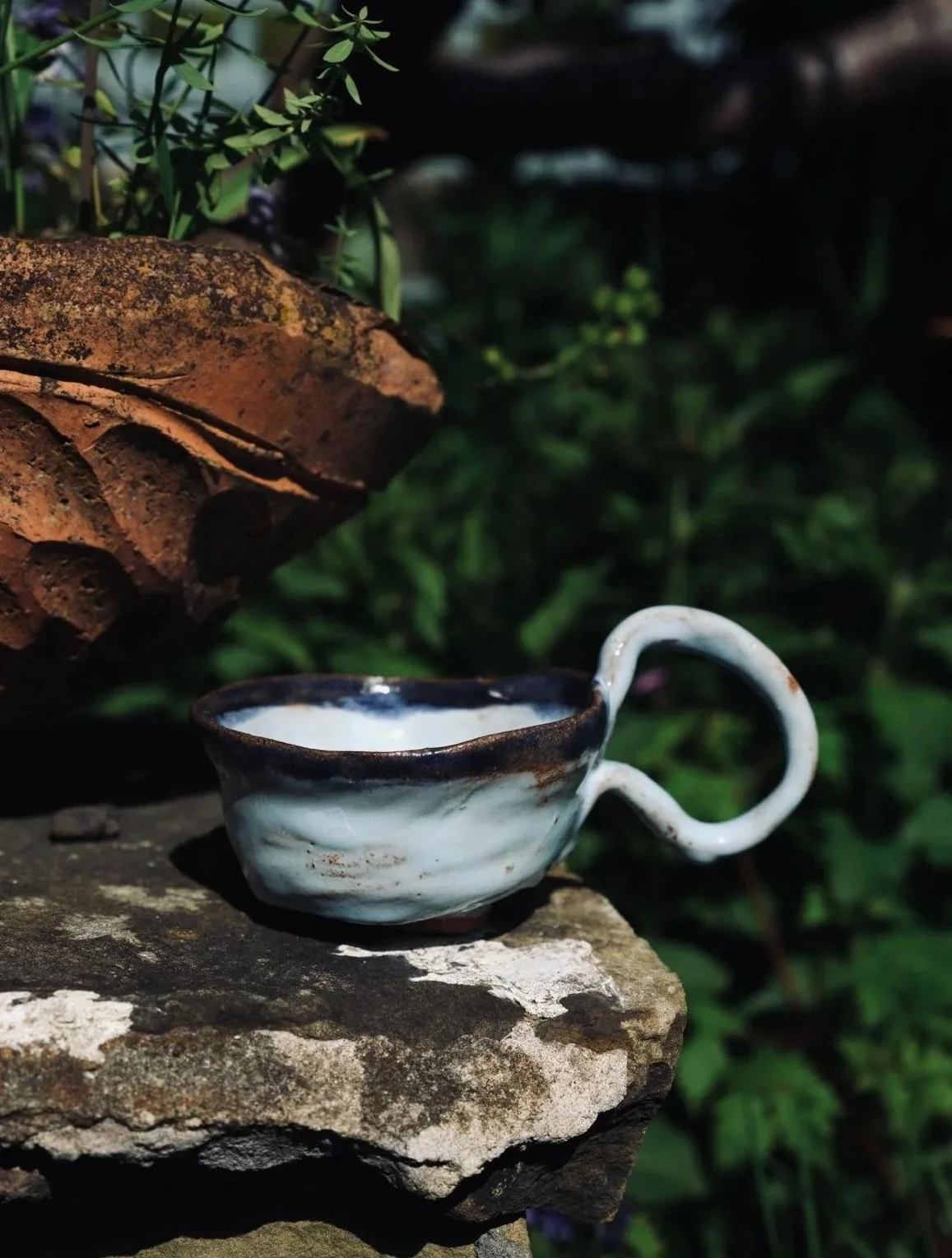 A ceramic teacup with a swirl handle on a rock, surrounded by greenery and stacked stones.