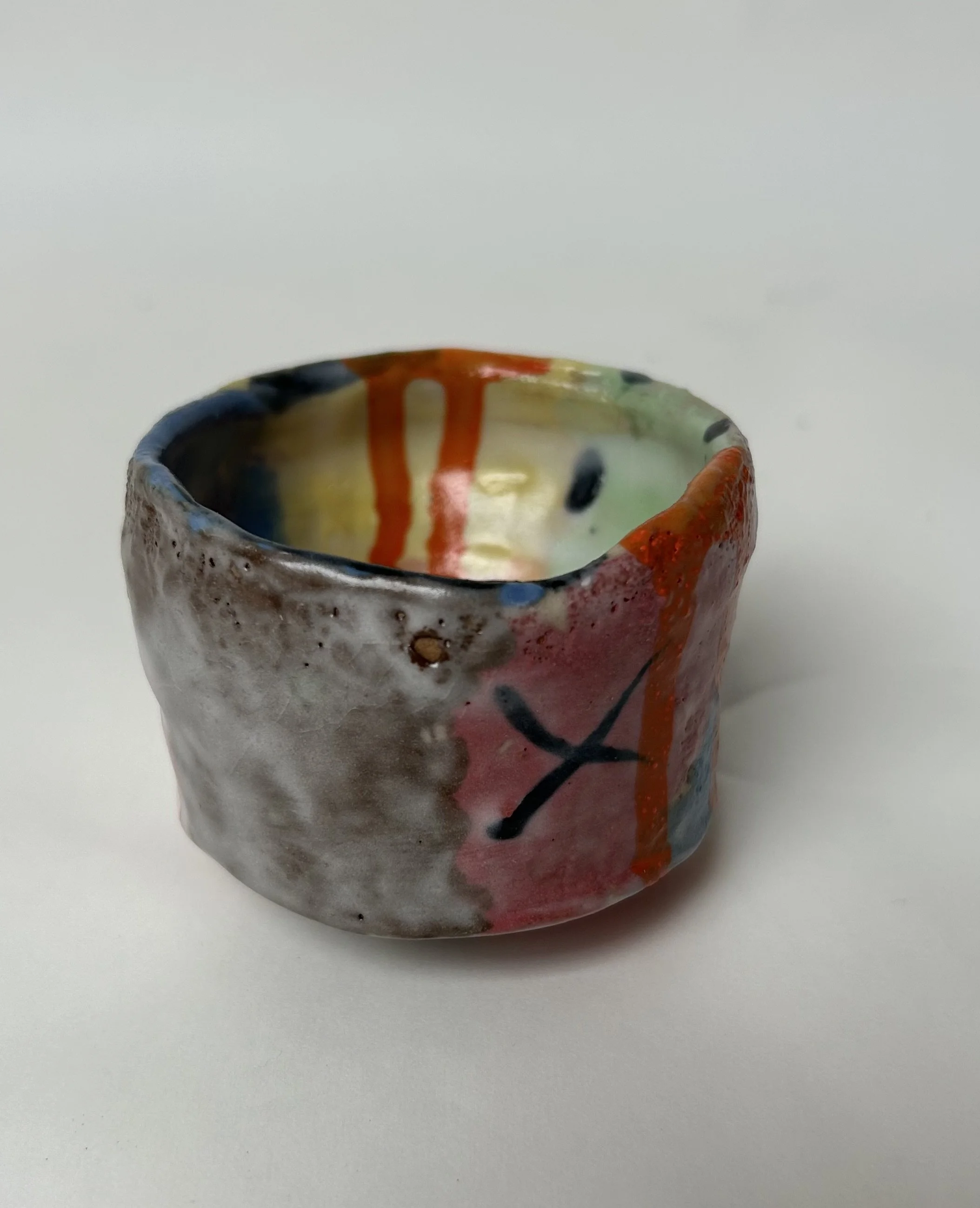 Close-up of a colorful handmade ceramic cup with abstract painted designs, set on a plain white background.