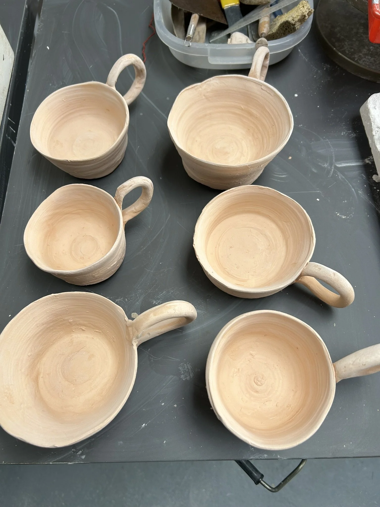 Unfinished ceramic cups with handles on a black work table, surrounded by pottery tools and supplies.