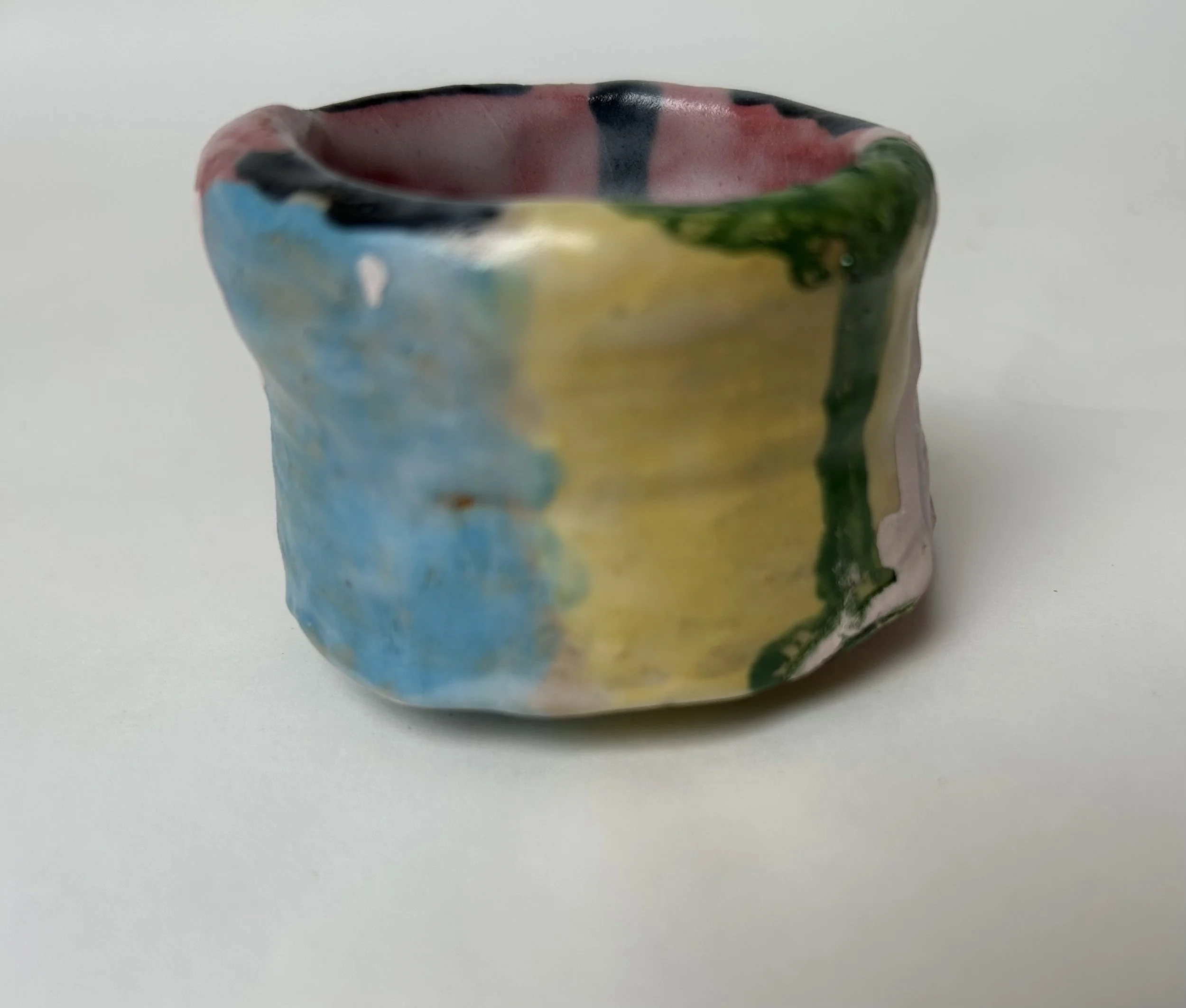 Close-up of a small, colorful ceramic bowl with a glossy finish, featuring a multicolored abstract pattern with red, green, blue, and yellow hues, set against a plain light background.