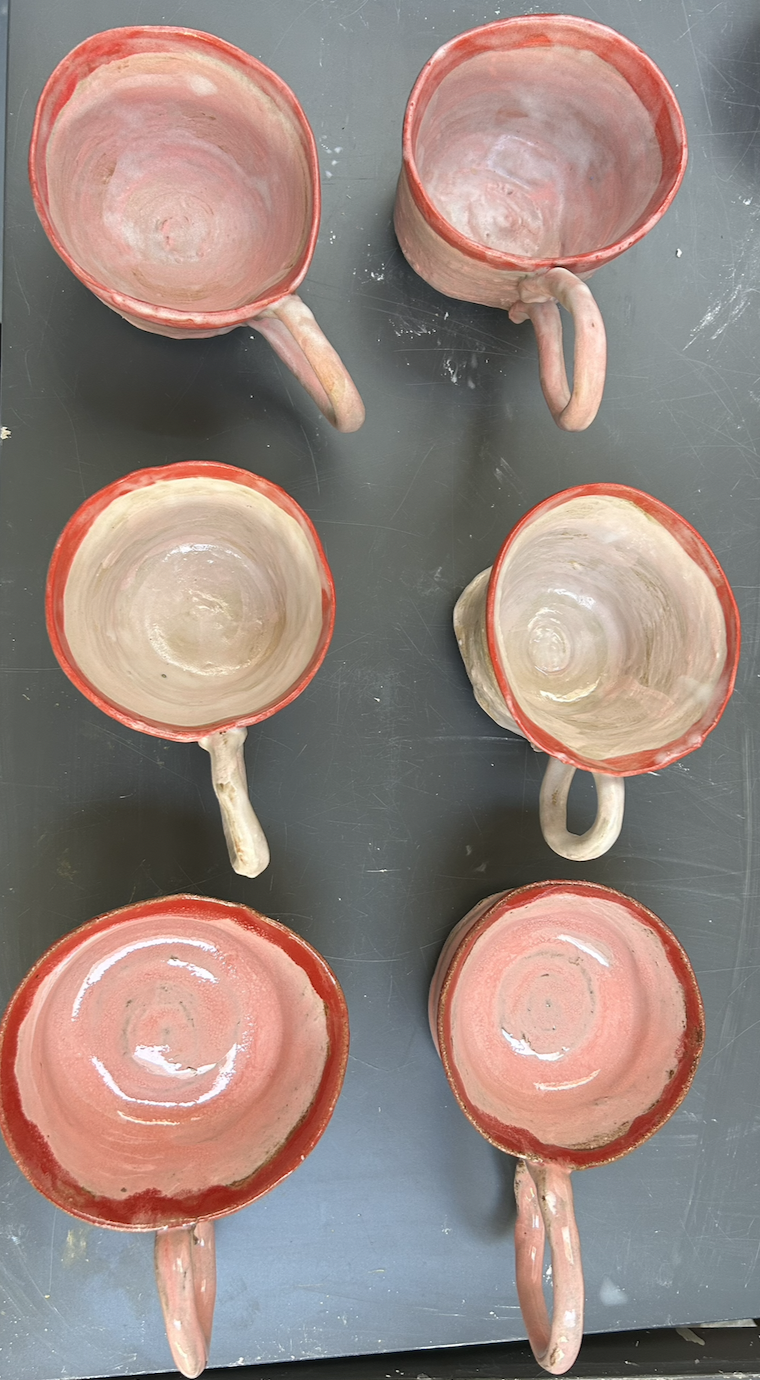 Six ceramic cups with handles, painted in shades of pink and red, arranged upside-down on a gray surface.