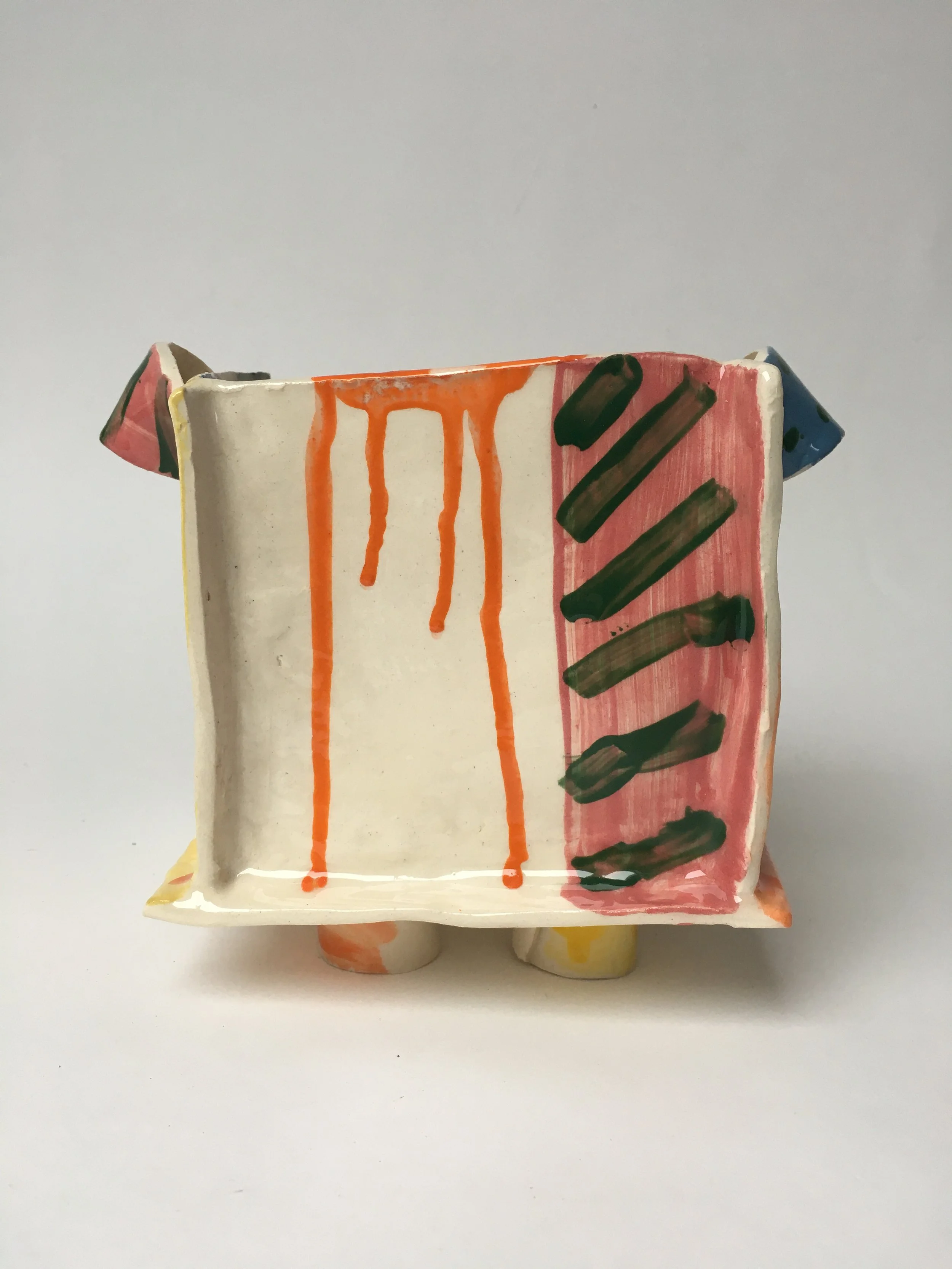 A ceramic sculpture of an abstract figure with painted stripes and drips, featuring a rectangular, box-like top and rounded bottom, placed on a flat surface against a plain background.