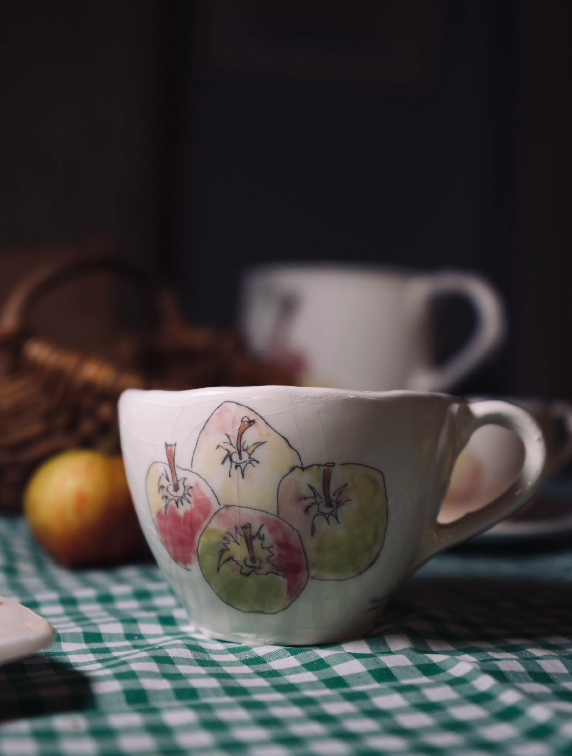 A teacup with a watercolor-style illustration of colorful apples and a handle, placed on a green and white checkered tablecloth, with blurred apples to the side and a dark background.