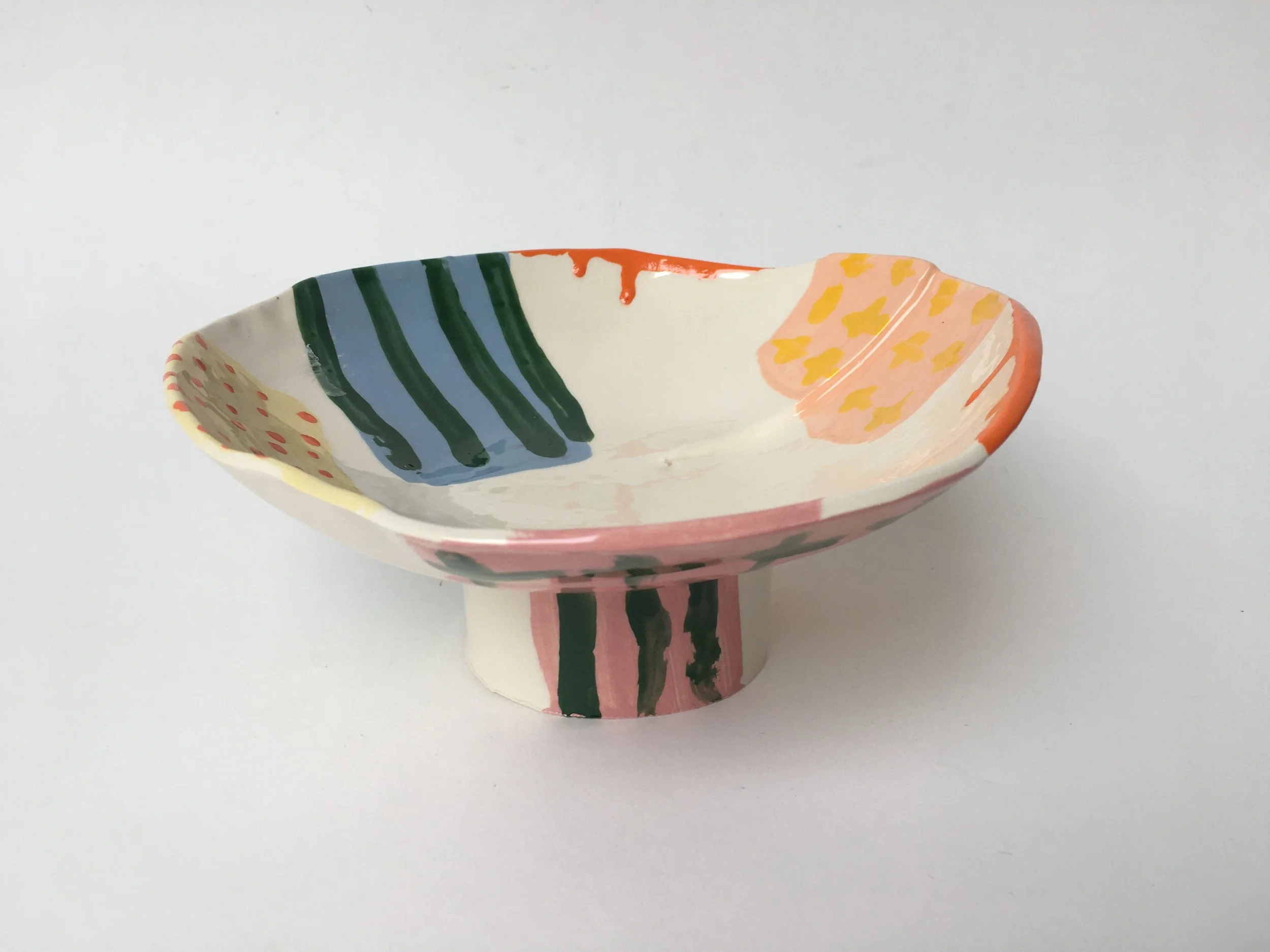 A decorative ceramic bowl with colorful painted patterns, including green stripes, yellow dots, and red accents, set against a plain white background.