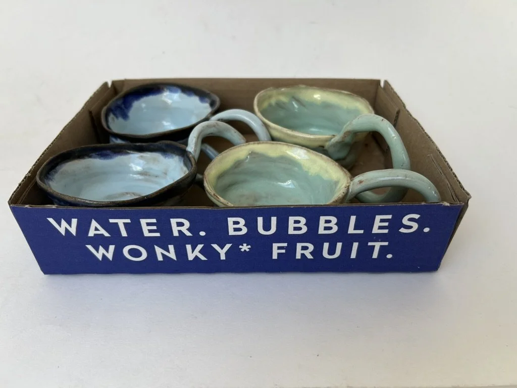 Six ceramic mugs with water-like glaze, inside a blue cardboard box labeled 'Water. Bubbles. Wonky* Fruit.'