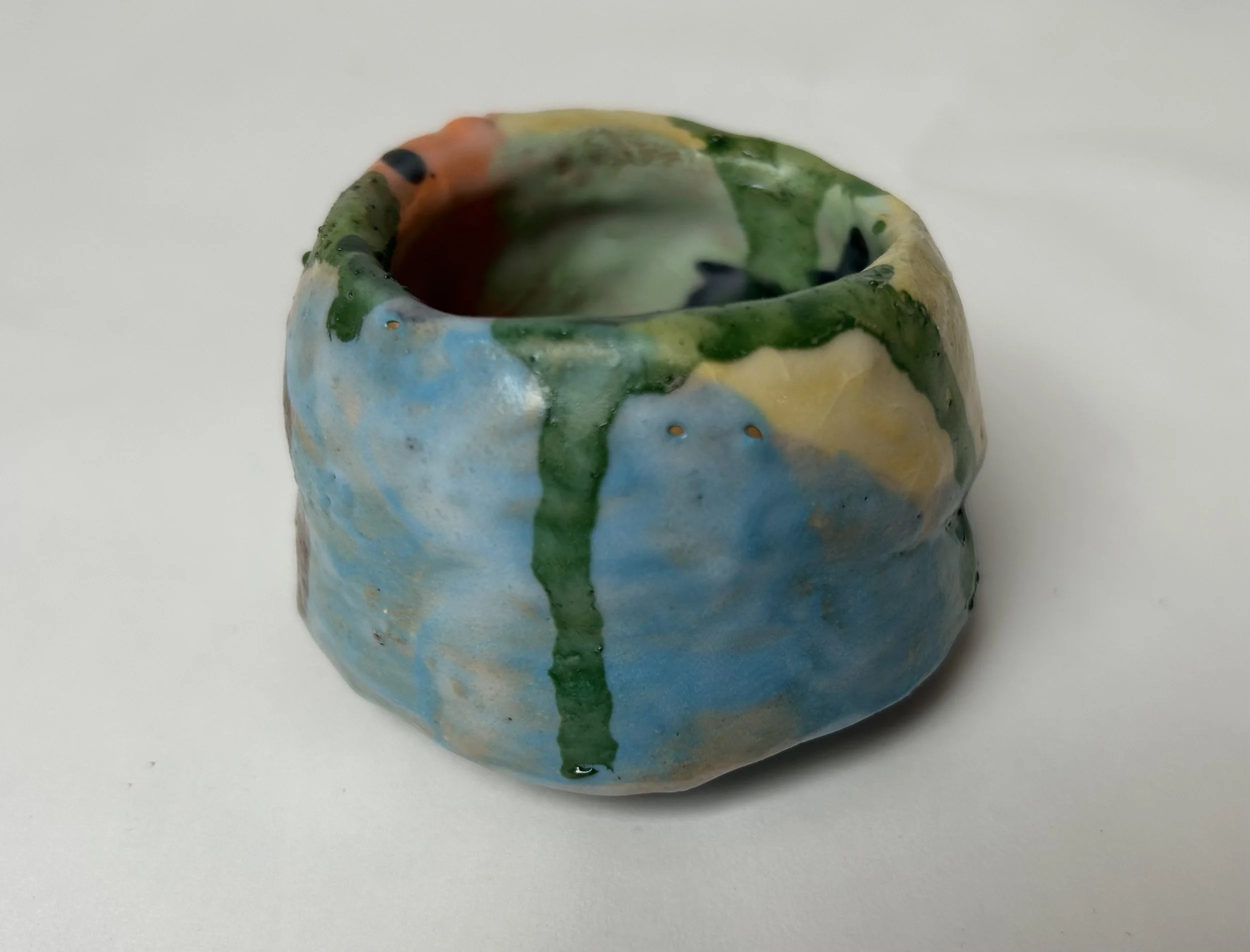A small ceramic cup with a colorful, abstract glaze pattern in green, blue, yellow, orange, and black, placed on a plain white surface.