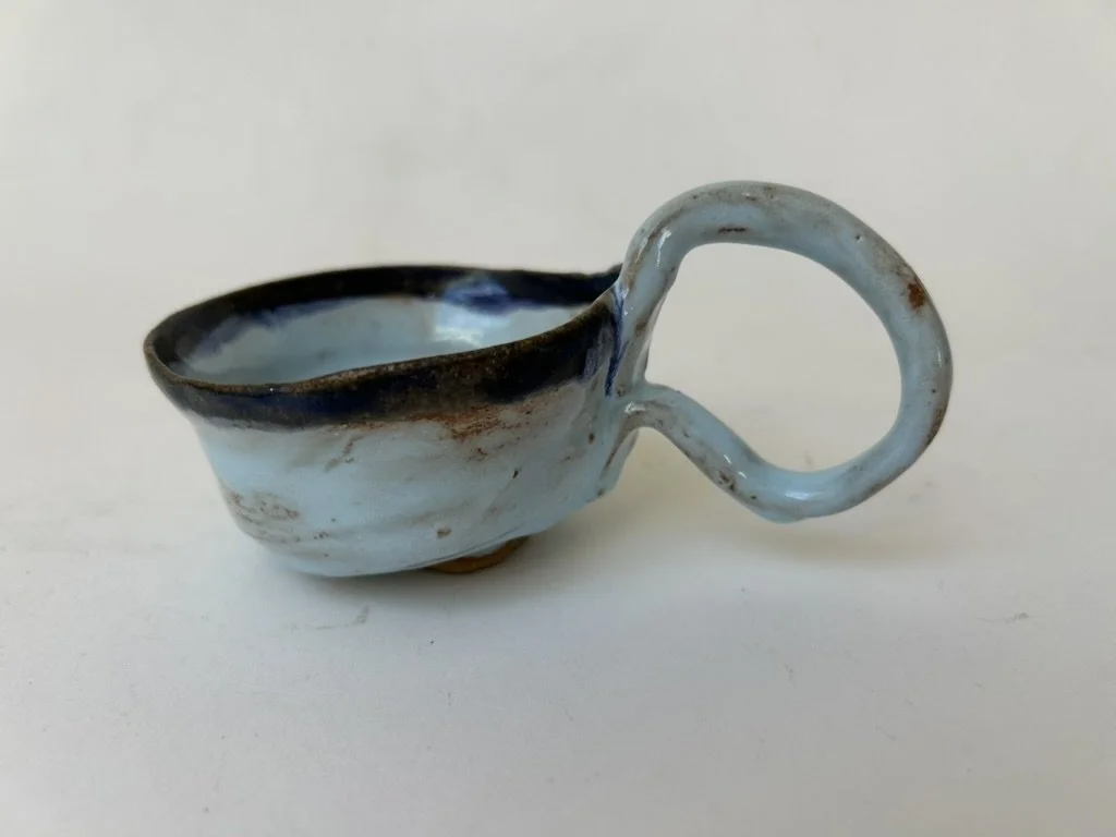 Handmade ceramic cup with a rough, rustic finish and a looped handle, glazed in shades of blue, white, and brown.