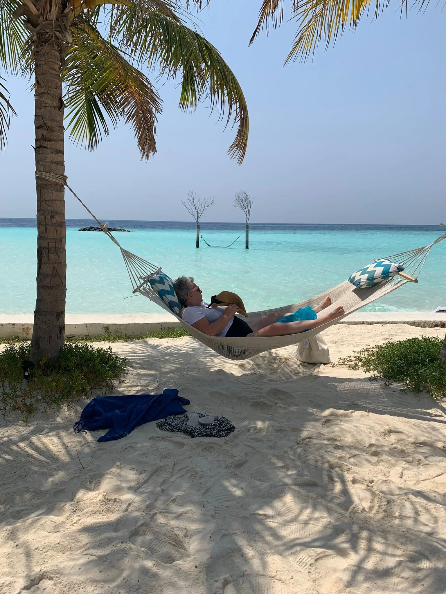 A woman relaxing on a hammock under a palm tree on a beach with white sand, turquoise water, and a clear blue sky.