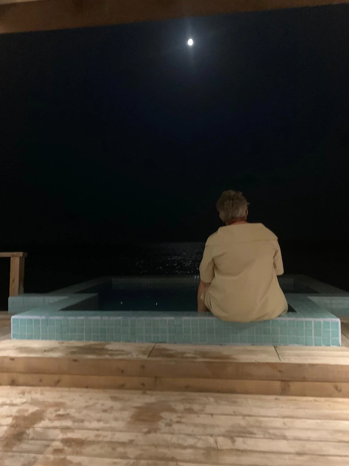 A woman sitting on the edge of a small pool at night, gazing at the bright moon in the sky over the water.