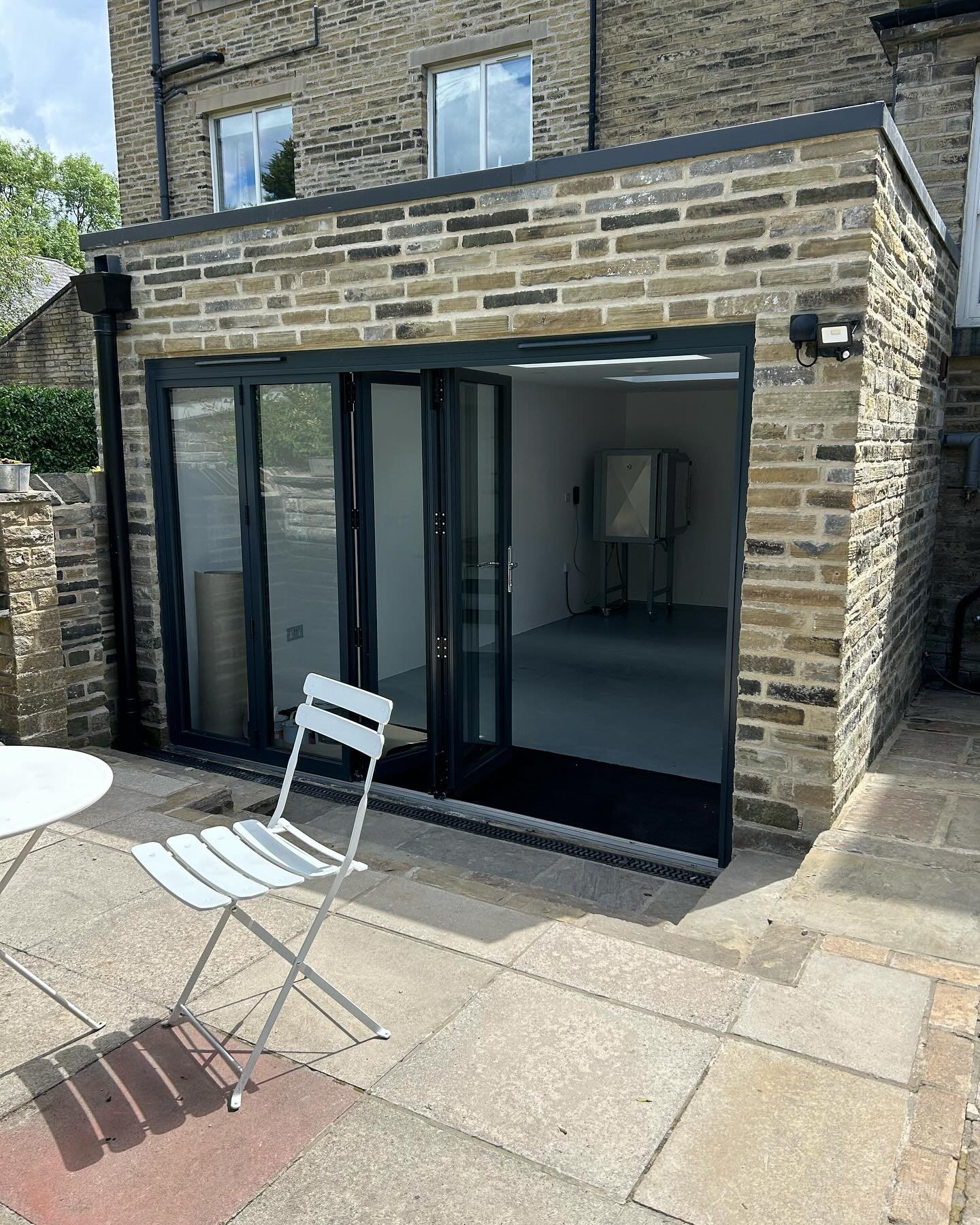 Outdoor patio area with a brick building having a glass-door extension, white outdoor chairs, a small round table, and potted plants, under a partly cloudy sky.
