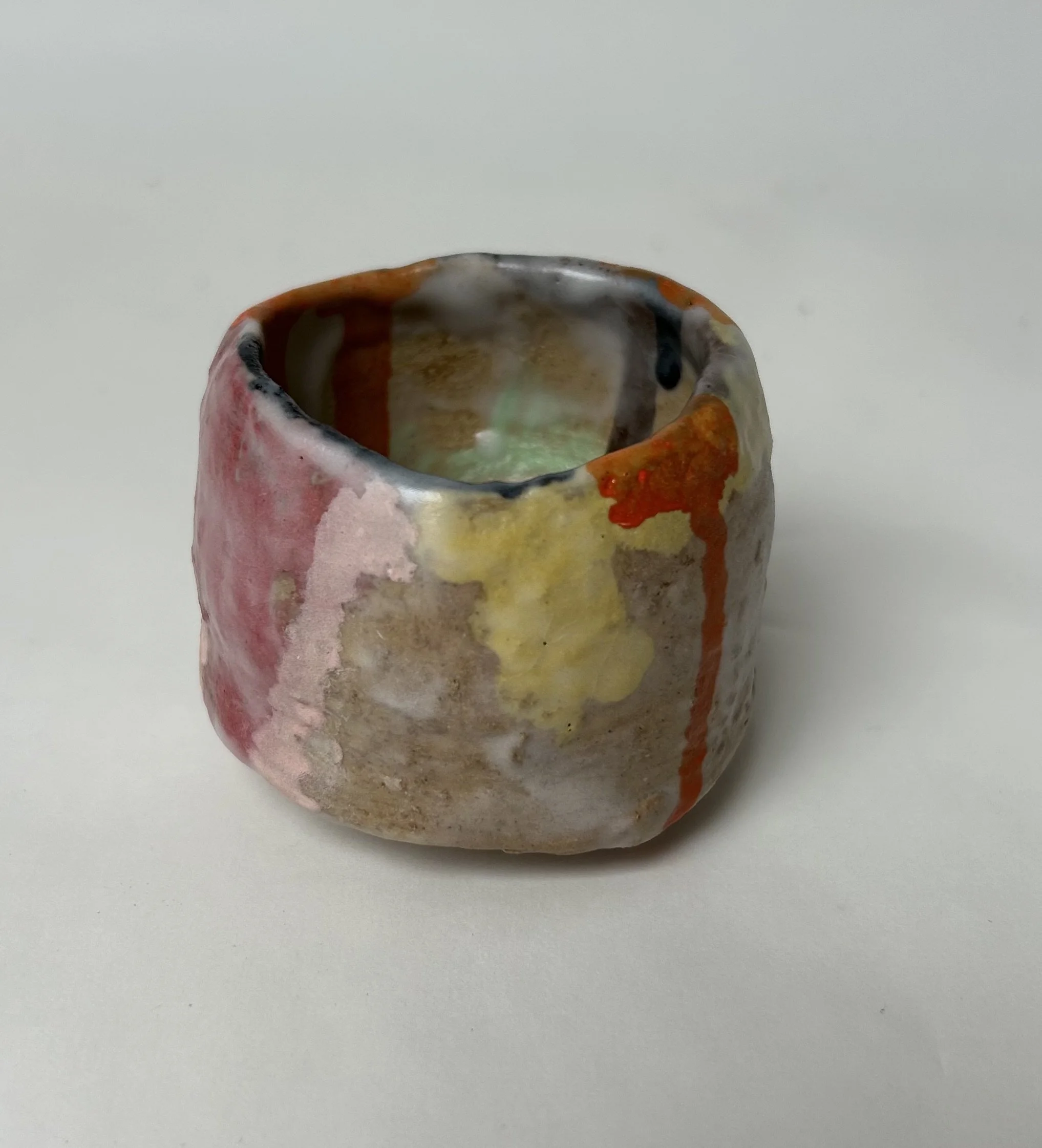 Close-up of a colorful ceramic cup or vessel with a rounded top, featuring a mix of pink, brown, yellow, and black glaze, set against a plain white background.