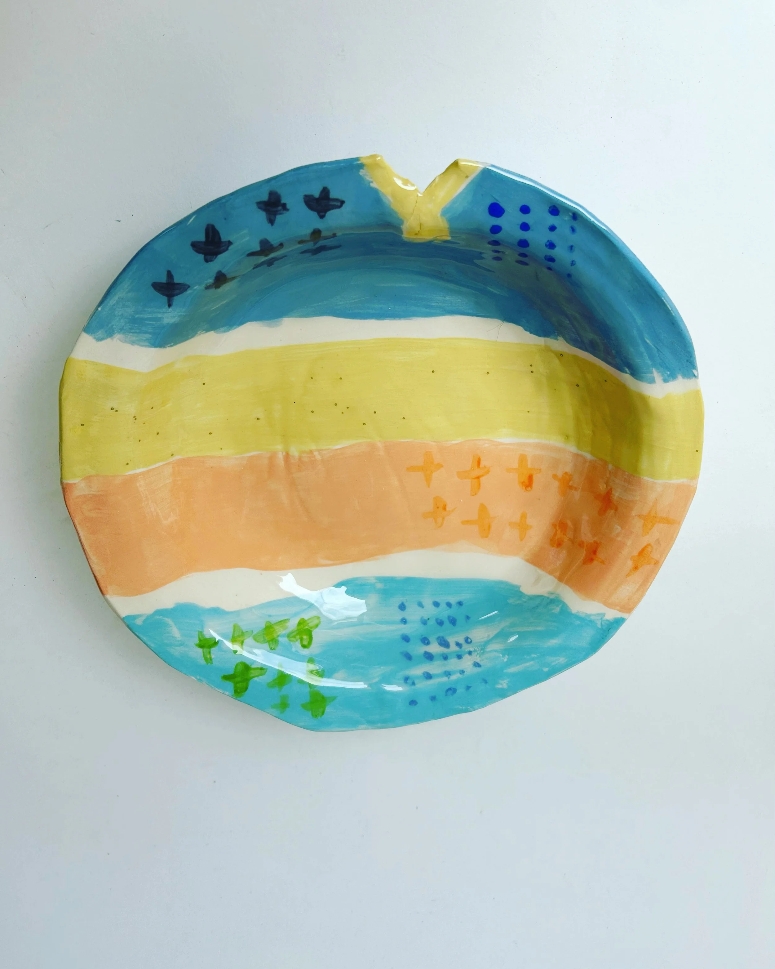 Colorful, hand-painted ceramic plate with abstract designs in stripes of blue, yellow, orange, and green, placed on a plain white surface.