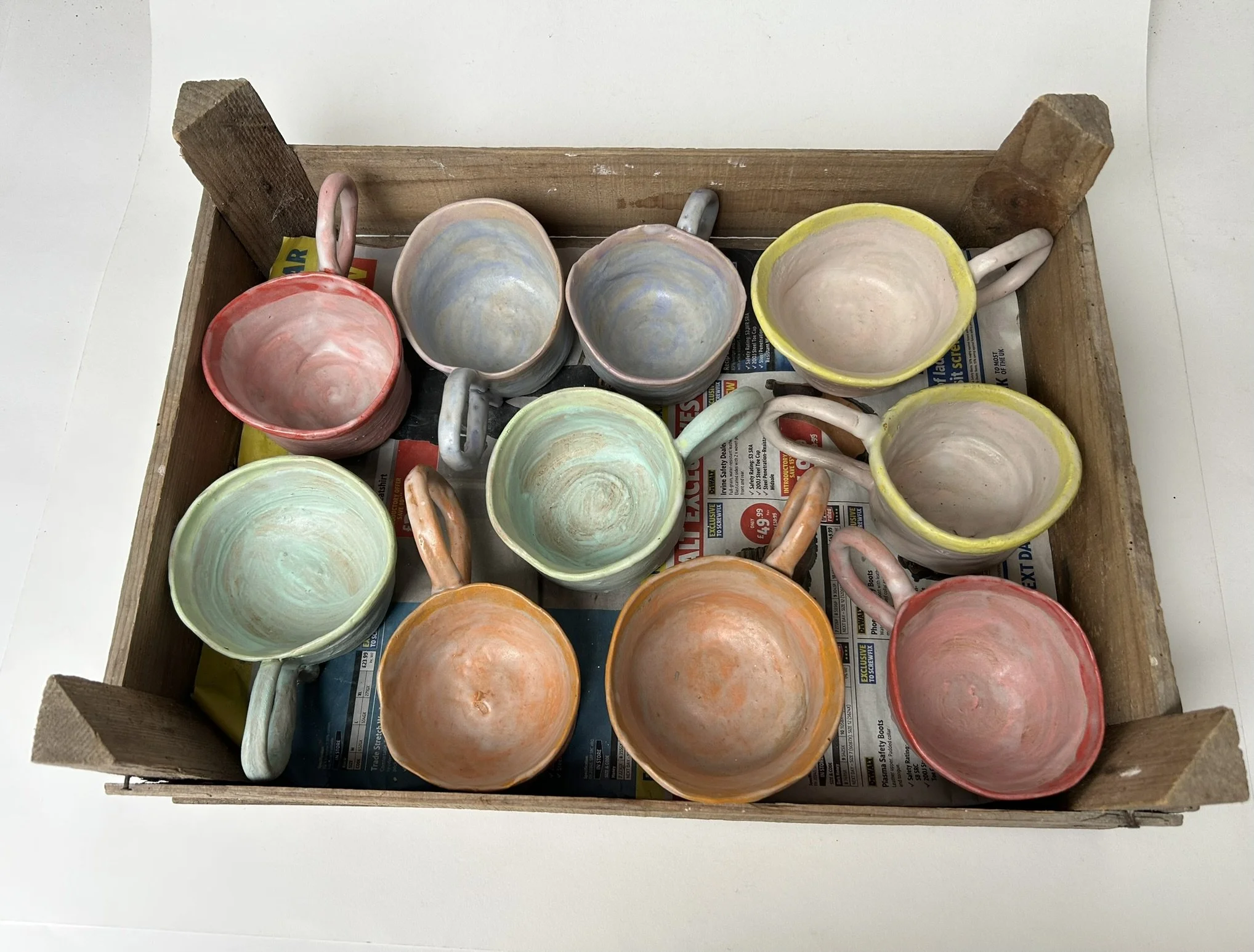 Colorful ceramic mugs with pastel and vibrant hues inside a wooden crate lined with newspaper.