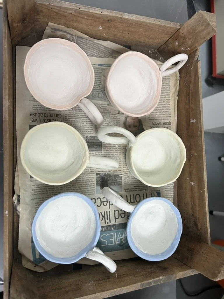 Six painted ceramic coffee mugs with painted handles, arranged in two columns of three on a wooden tray covered with newspaper.