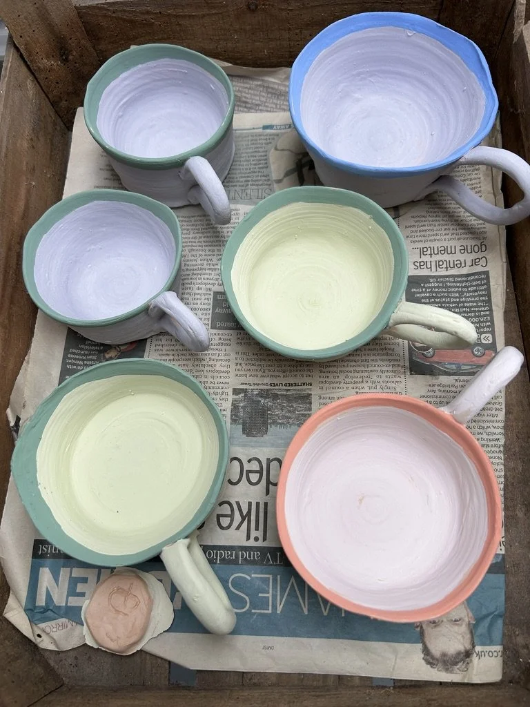 Six ceramic mugs and bowls in pastel colors on newspaper, waiting to be glazed or painted.